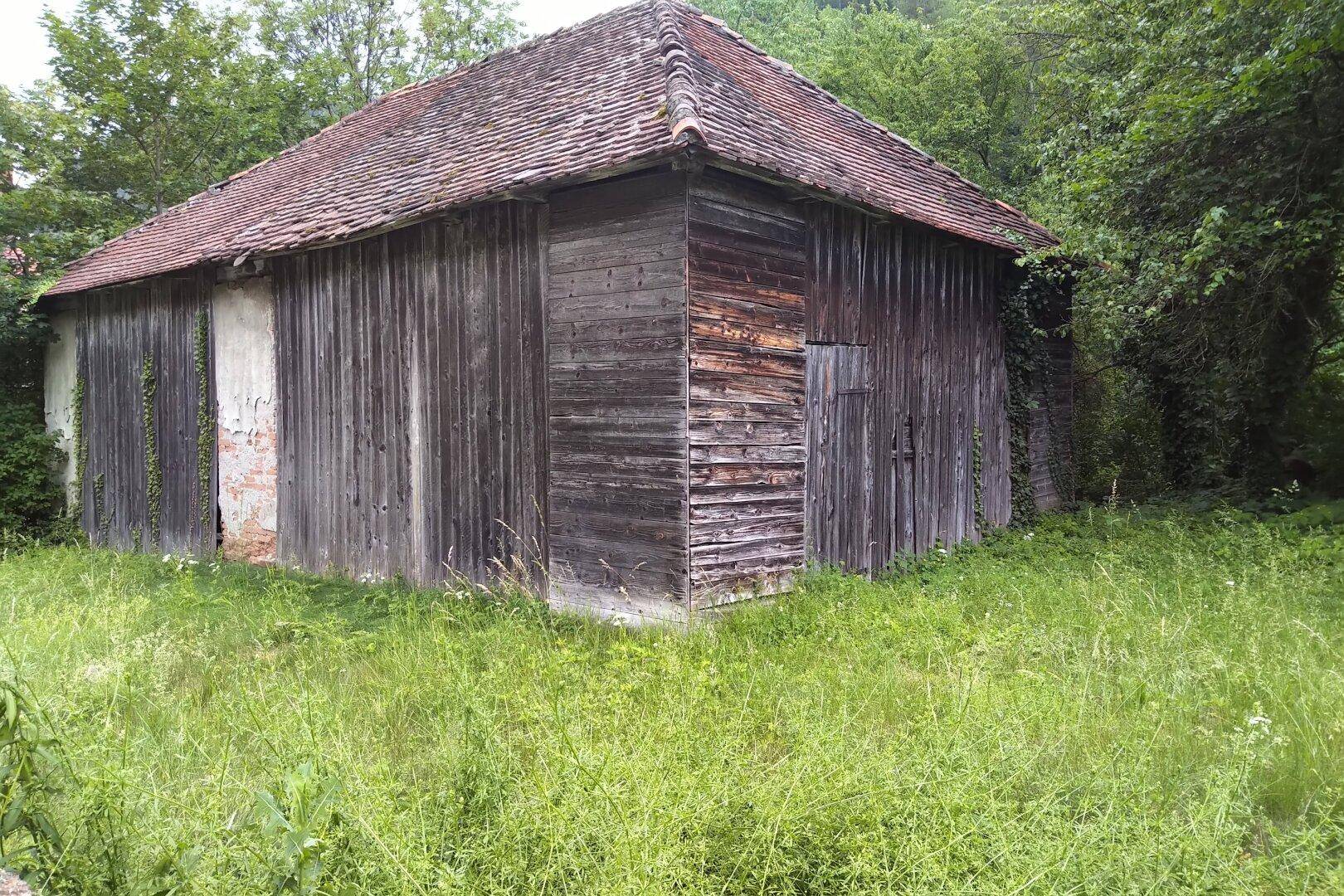 Rustikales Holzgebäude mit verwitterten Fassaden und Ziegeldach, umgeben von hohem Gras.