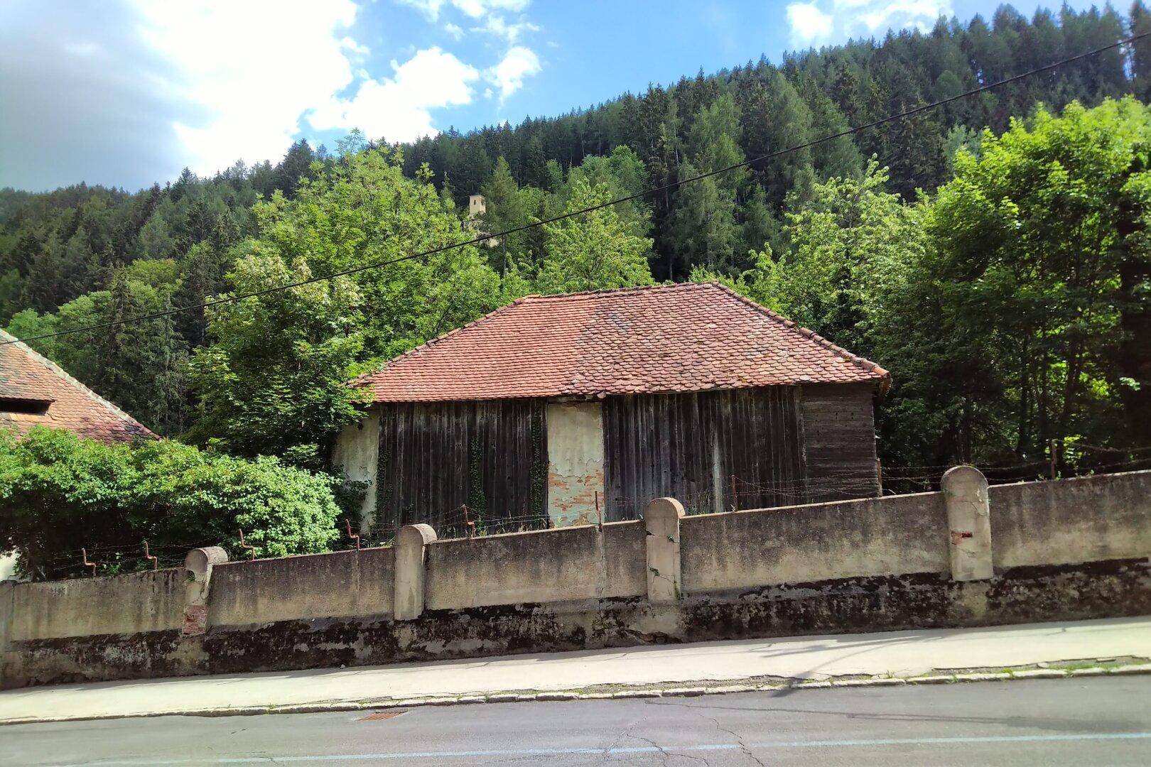 Altes Holzgebäude mit Ziegeldach und Steinmauer, umgeben von Bäumen und Grün.