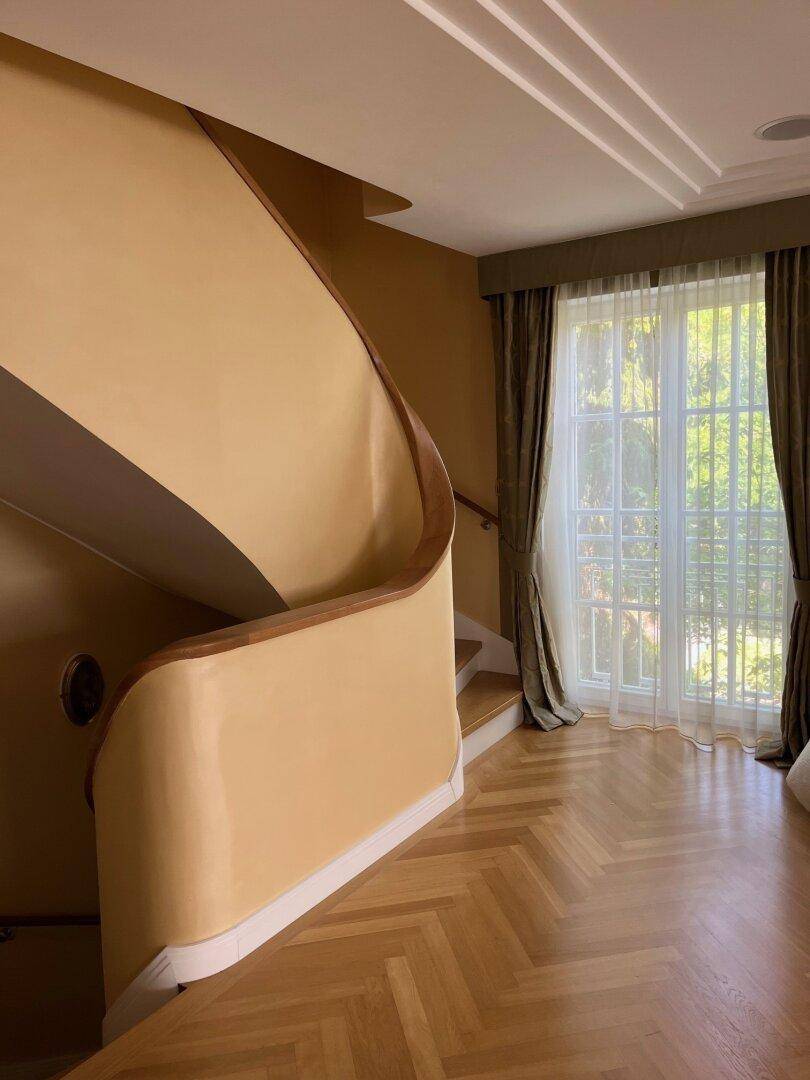 Heller Flur mit geschwungener Treppe, Fischgrätparkett und großen Fenstern.