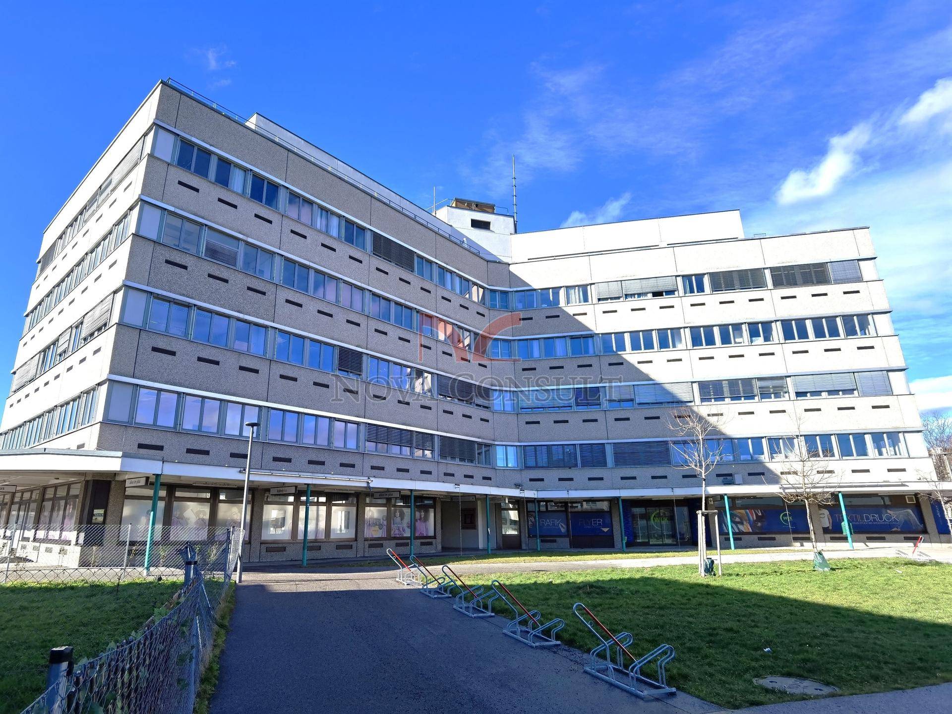 Modernes Bürogebäude mit vielen Fenstern und einer gepflegten Grünfläche davor unter blauem Himmel.
