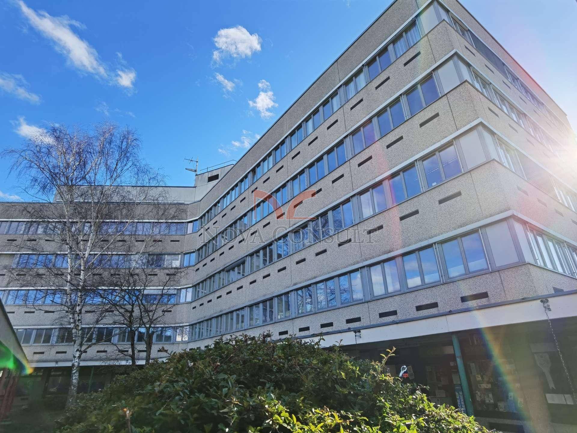 Großes Bürogebäude mit vielen Fenstern und einer modernen Fassade, umgeben von Bäumen und Sträuchern.