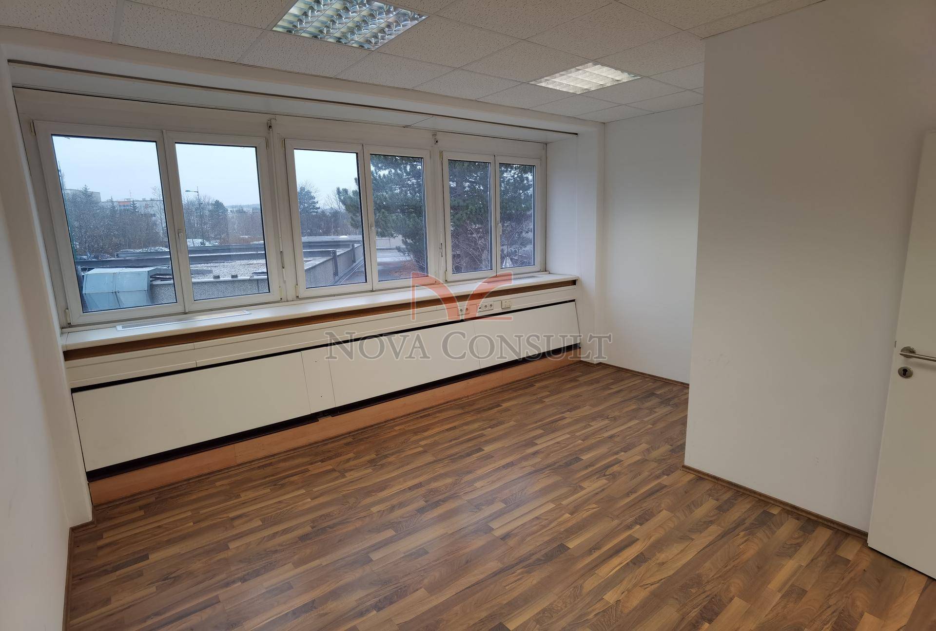 Helles Büro mit Holzboden und einer Fensterreihe, die einen Blick auf die Umgebung und Bäume bietet.