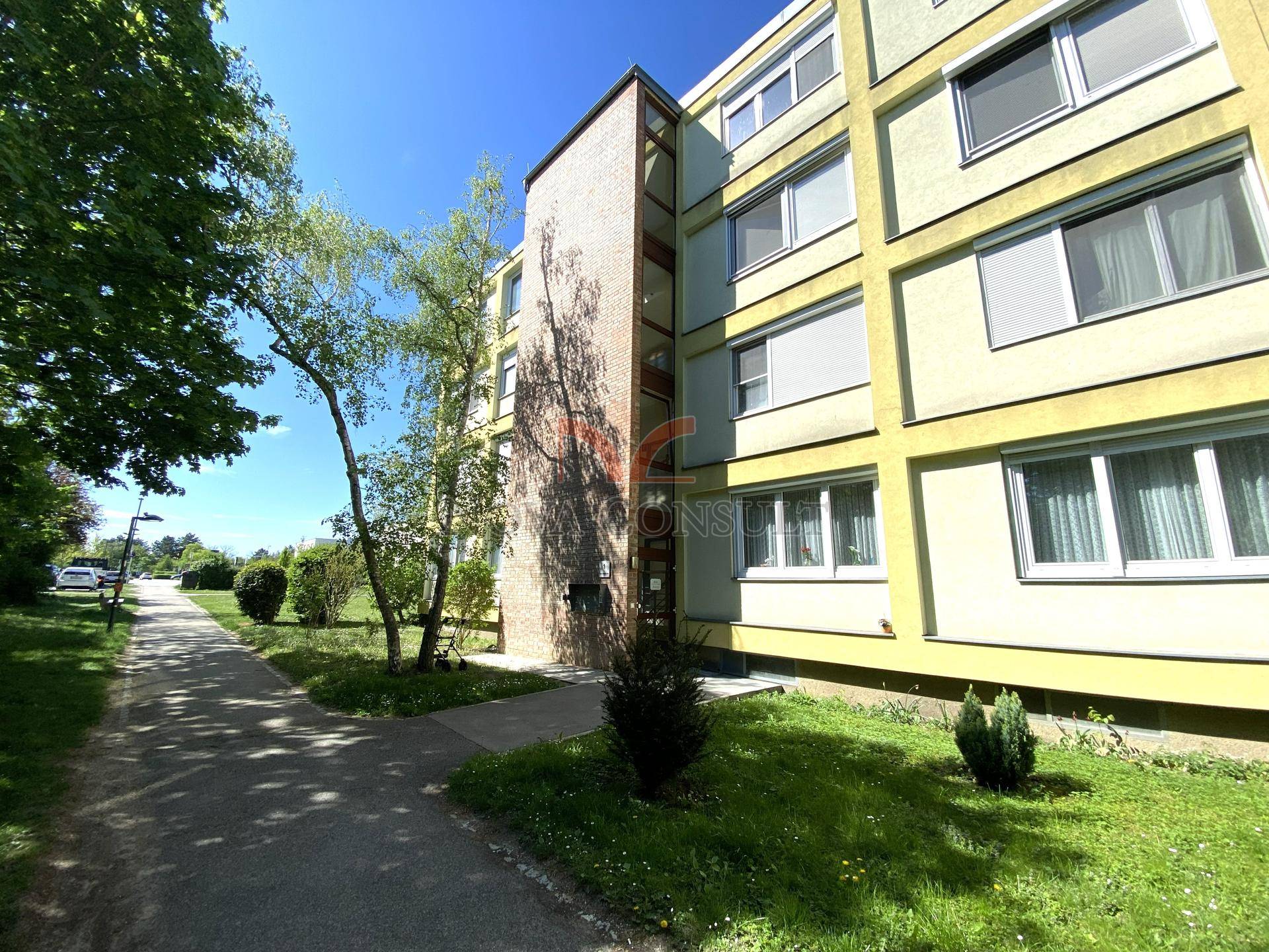 Eingangsbereich eines Wohngebäudes mit gelber Fassade, umgeben von gepflegten Grünflächen und Wegen.