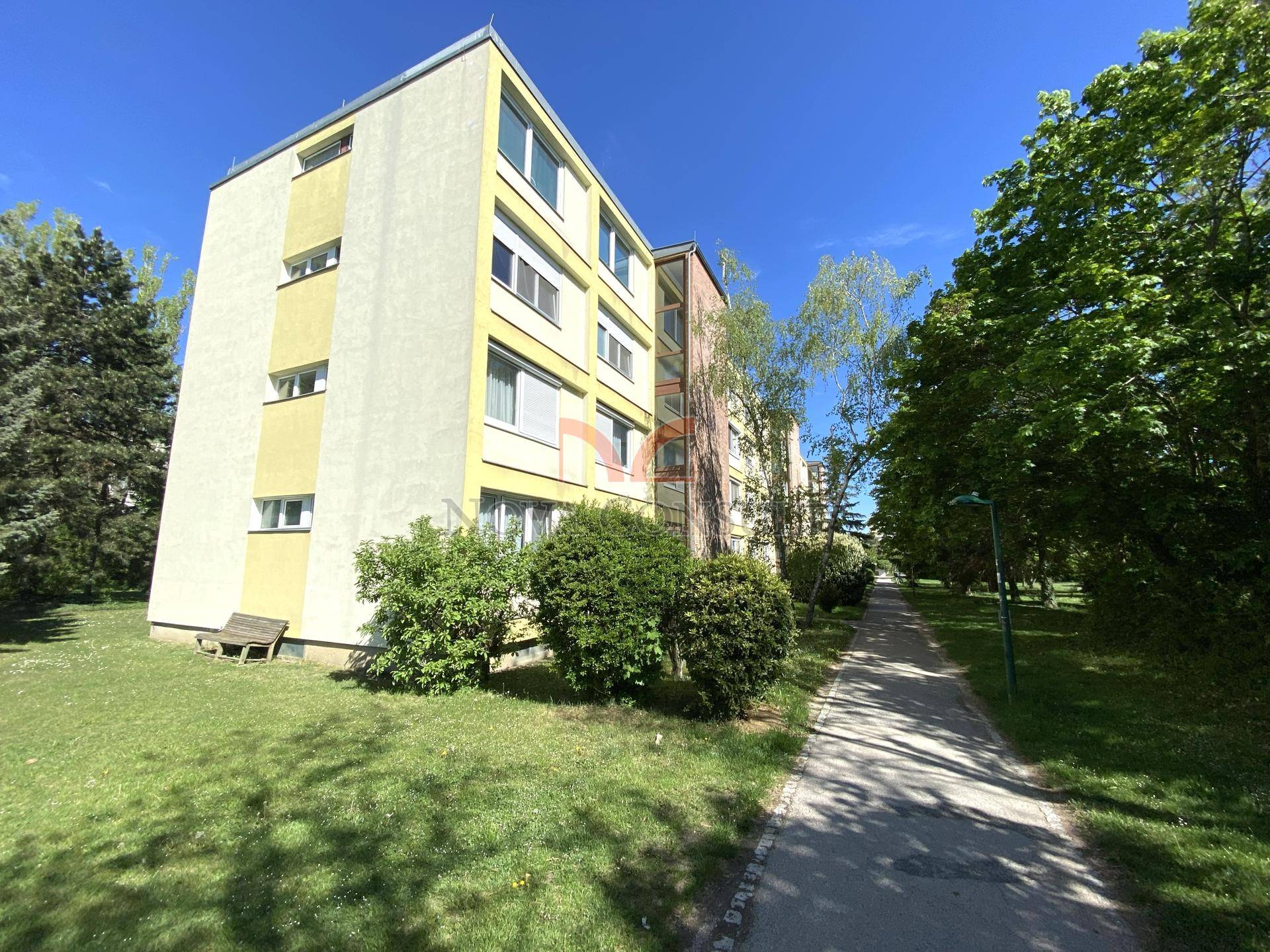 Mehrstöckiges Wohngebäude mit gelber Fassade, umgeben von Grünflächen und Bäumen.