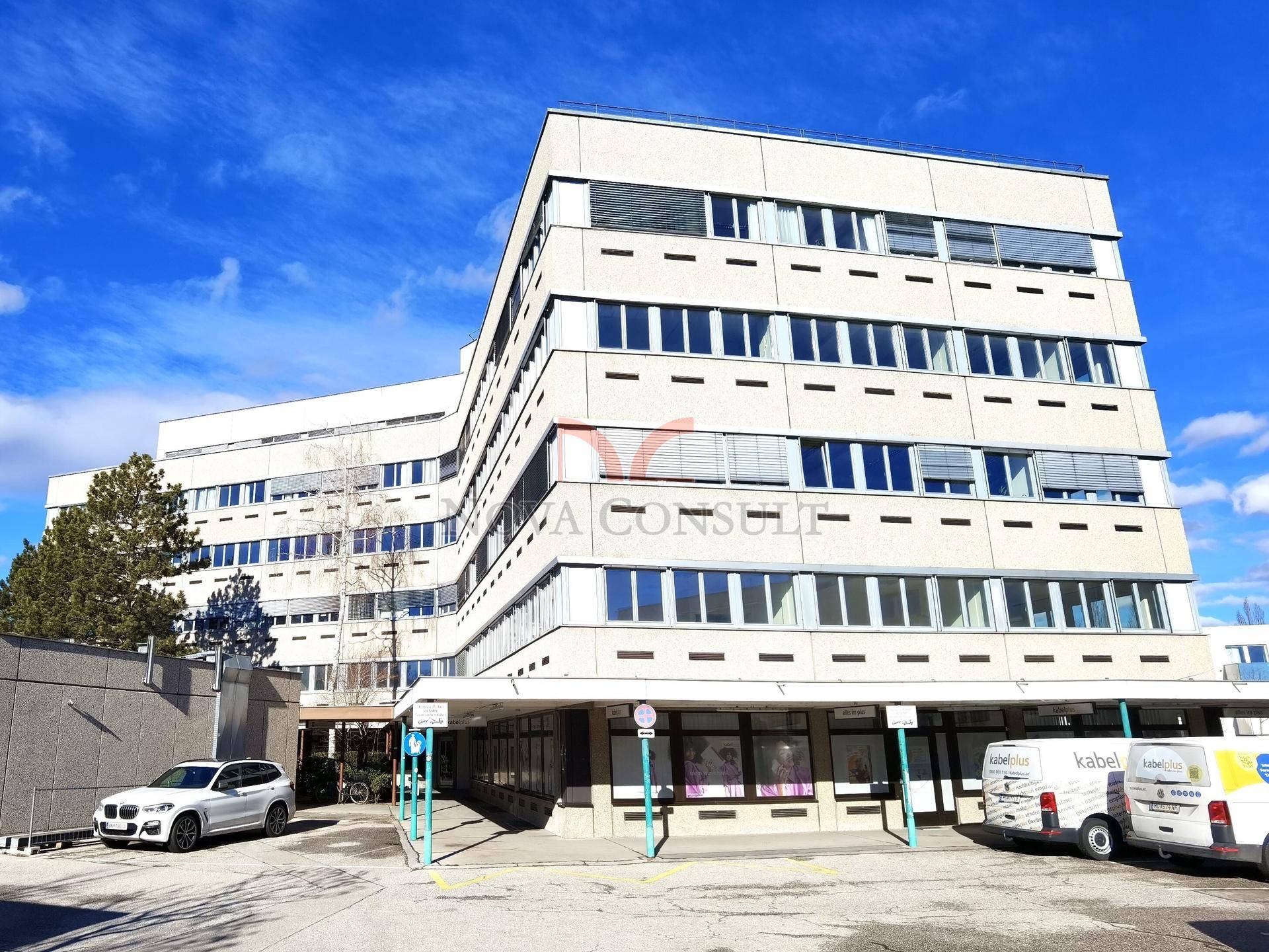Modernes Geschäftsgebäude mit mehreren Etagen, großen Fensterfronten und einem Parkplatz davor unter blauem Himmel.