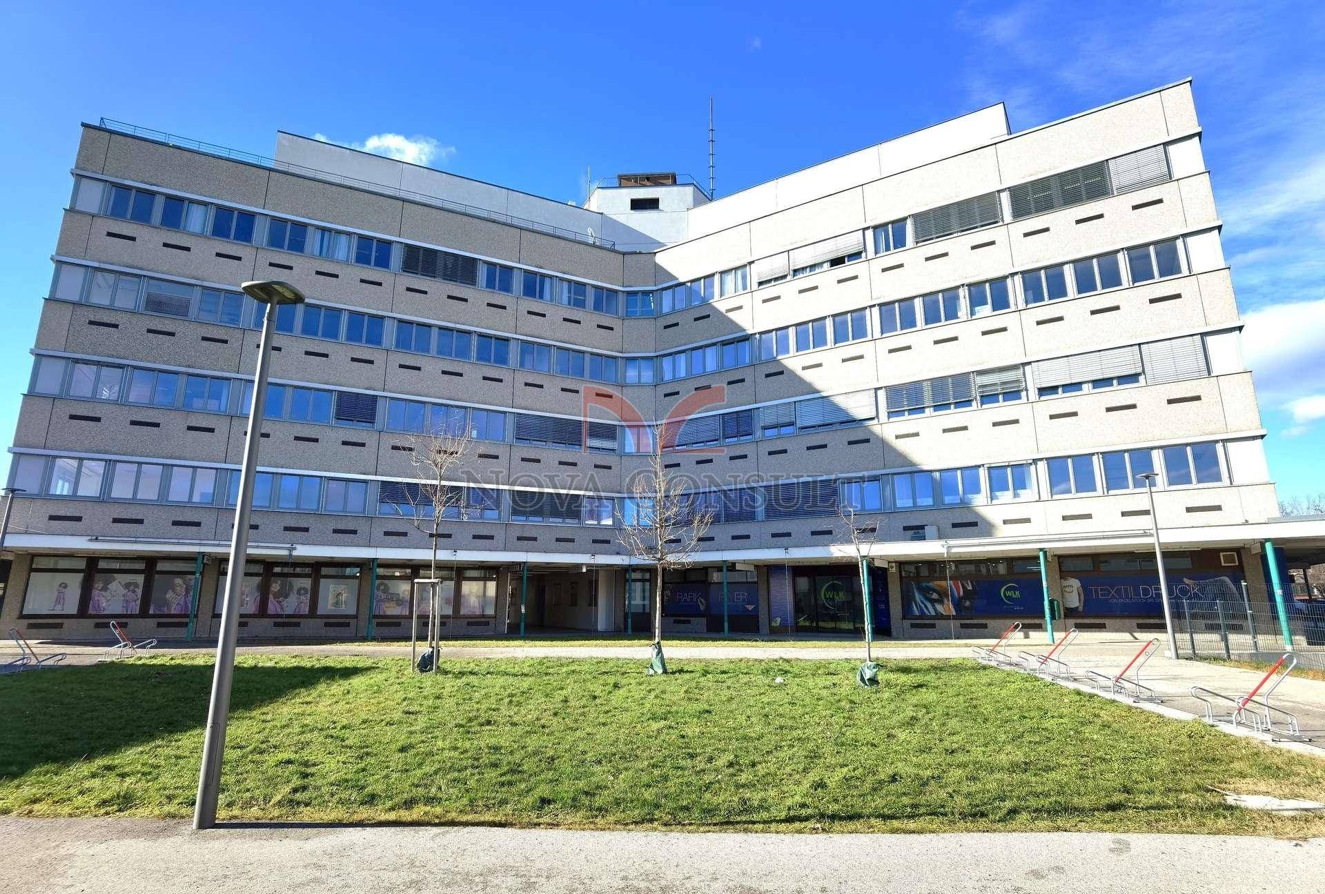 Weitläufiges Geschäftsgebäude mit Grünfläche und Geschäften im Erdgeschoss unter hellem, bewölktem Himmel.