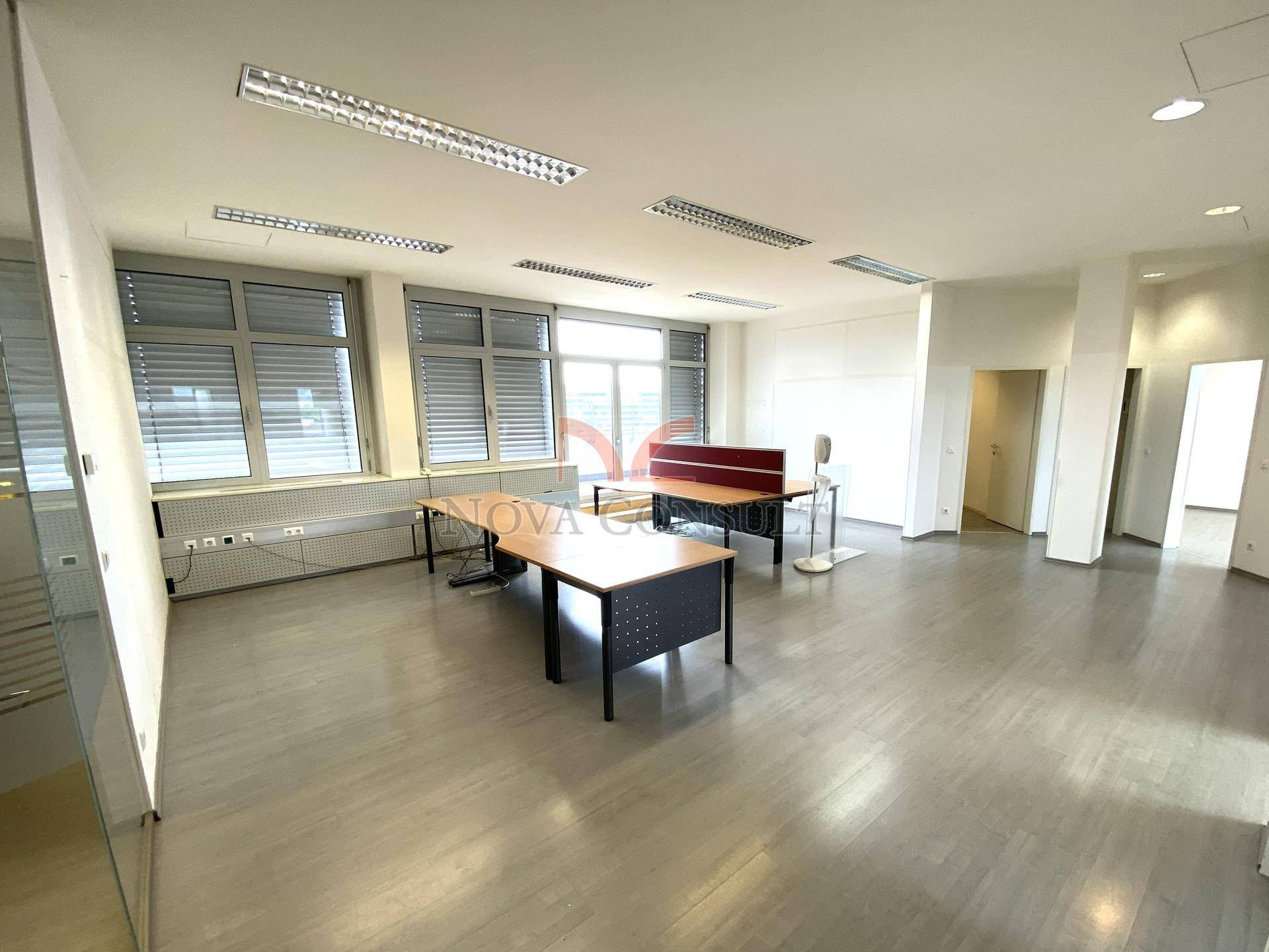 Weitläufiges Büro mit mehreren Arbeitsbereichen, großen Fenstern und hellem Laminatboden.