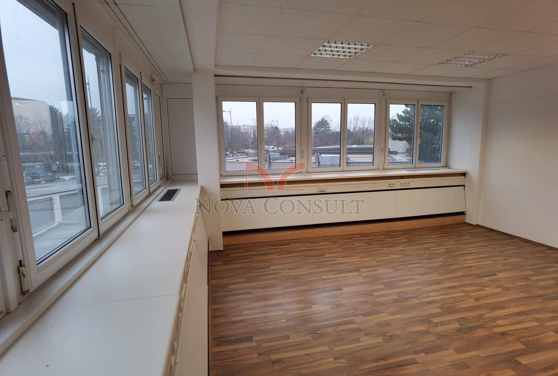 Geräumiges Büro mit Holzboden und zwei Fensterfronten, die den Raum mit viel natürlichem Licht durchfluten.