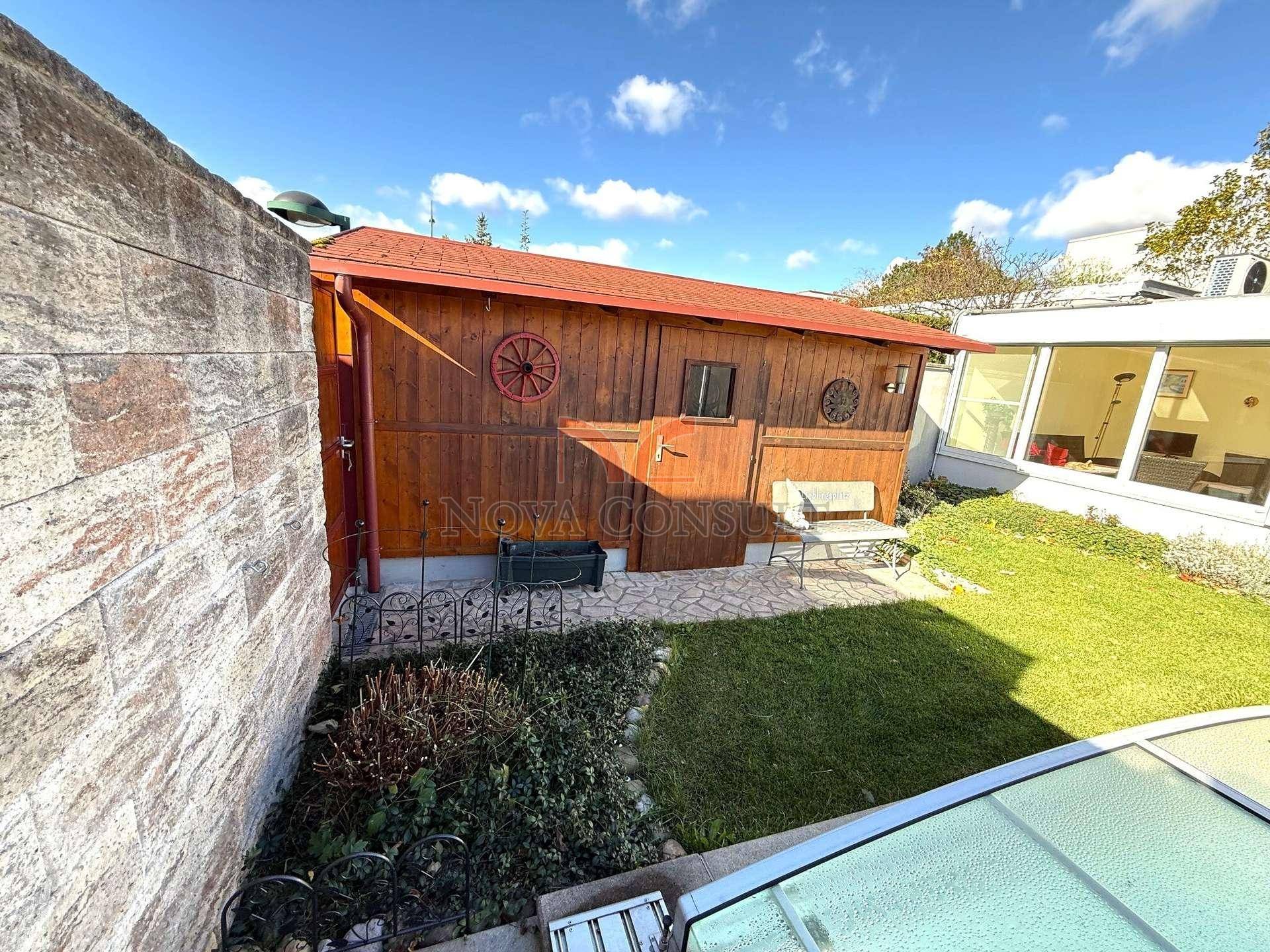 Rustikaler Holzschuppen im Garten, umgeben von Grünflächen und einer Steinmauer.