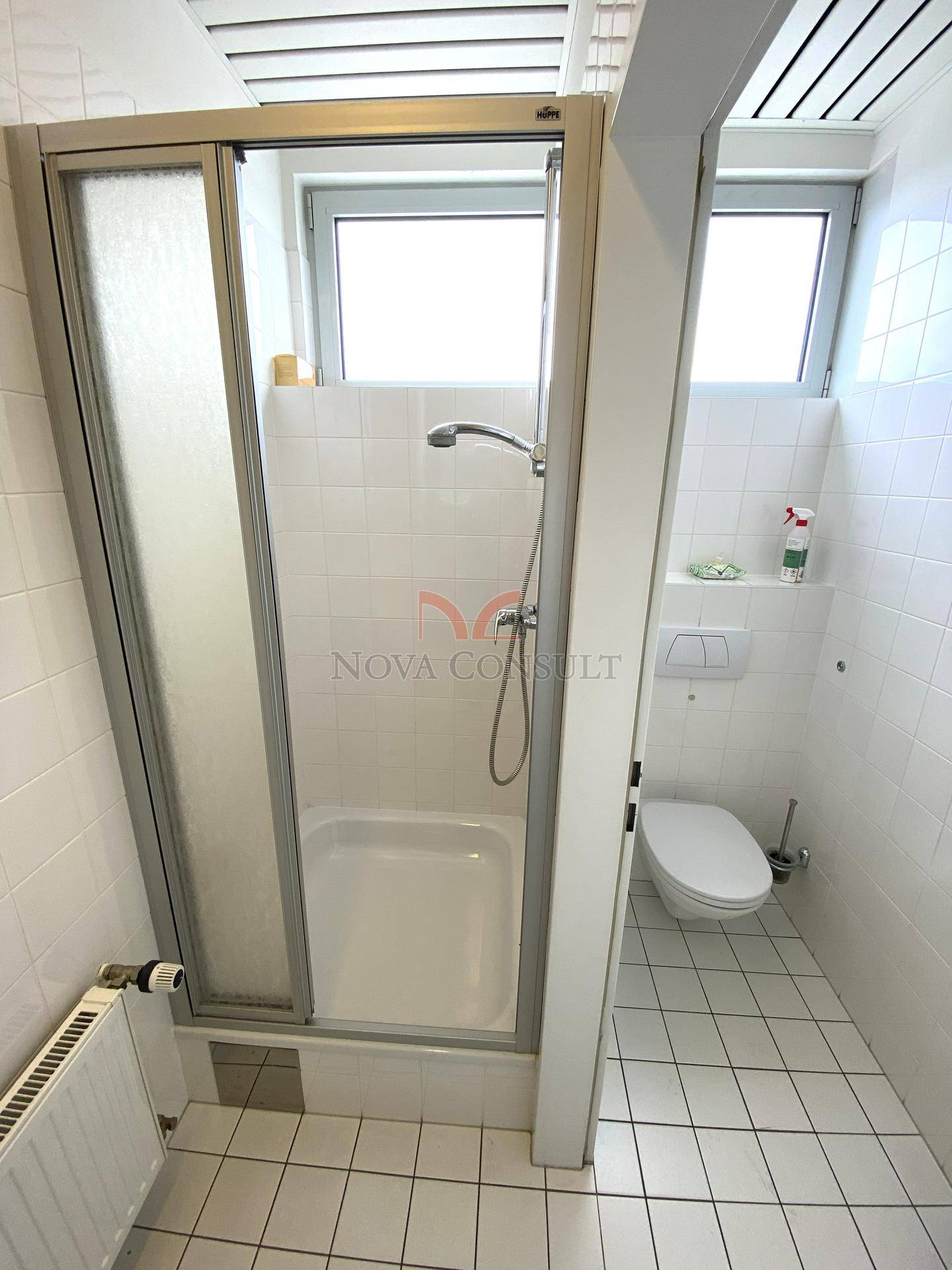Badezimmer mit Duschkabine, Toilette und Fenster, komplett gefliest in Weiß.
