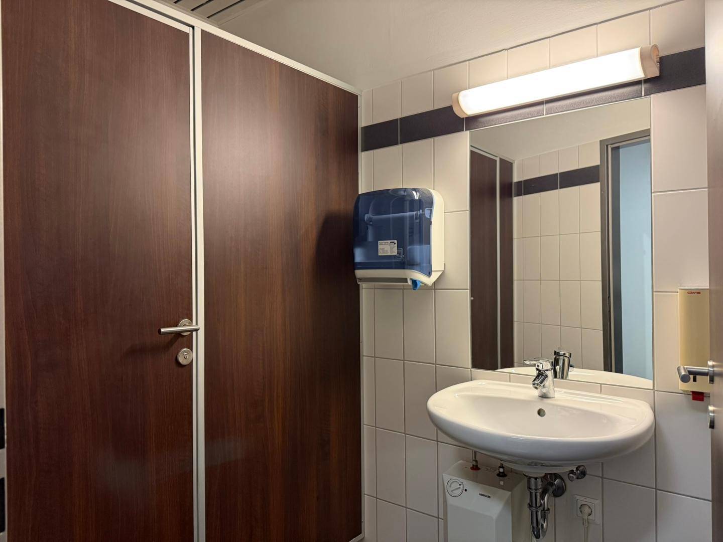 Funktionale Toilette mit Waschbecken, Spiegel und weißen Fliesen, ausgestattet mit einem Handtuchspender.