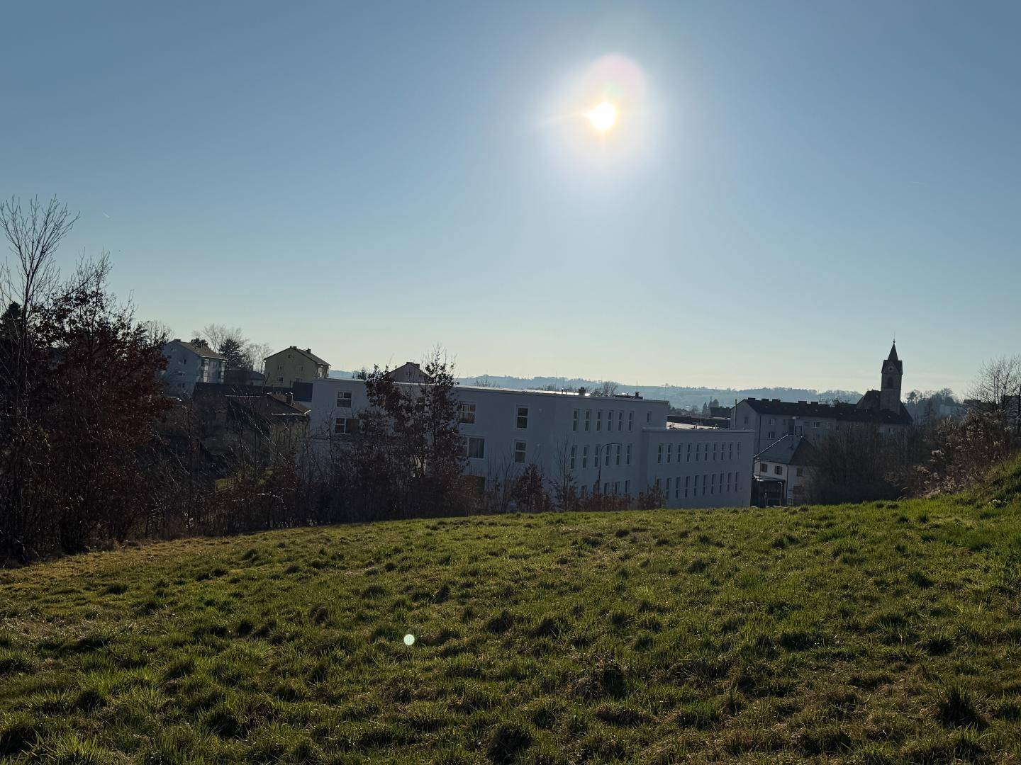 Weitläufige grüne Wiese unter strahlend blauem Himmel mit Gebäuden und einem Kirchturm in der Ferne.