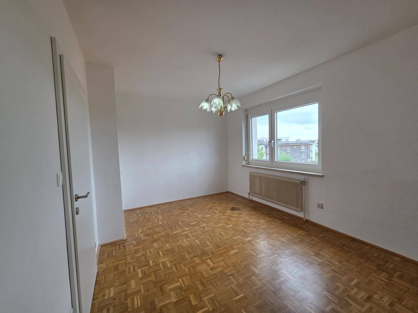 Geräumiges, unmöbliertes Zimmer mit Fischgrätparkett und großem Fenster.