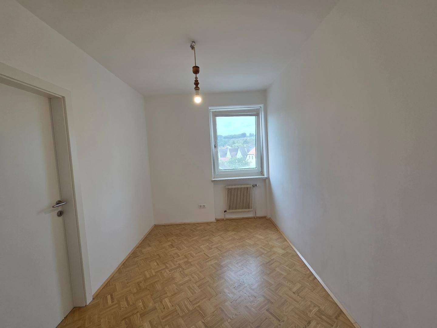 Kleineres, unmöbliertes Zimmer mit Parkettboden und Fenster mit Blick ins Grüne.