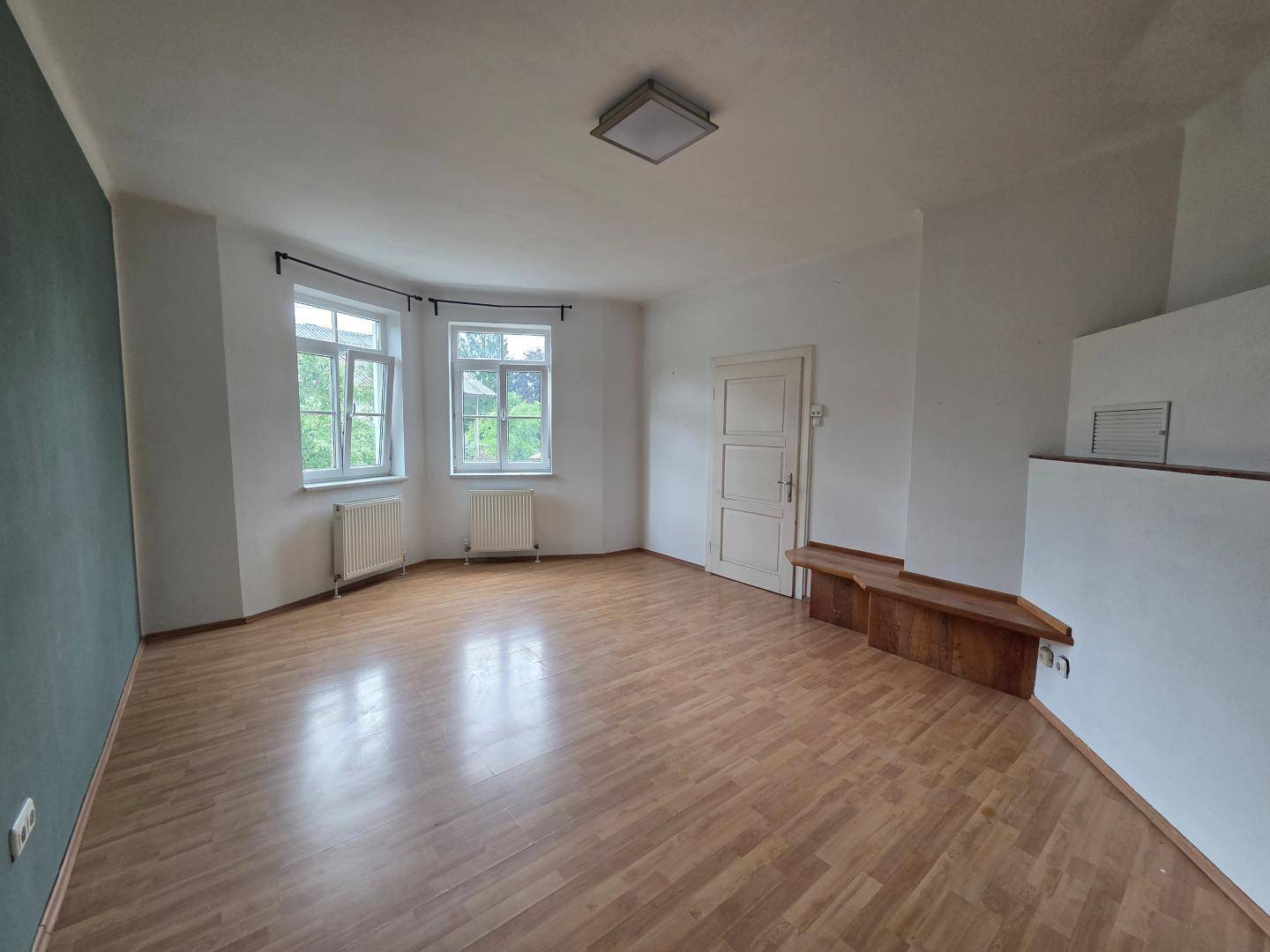 Helles, leeres Wohnzimmer mit Parkettboden, großen Fenstern und einer Nische.