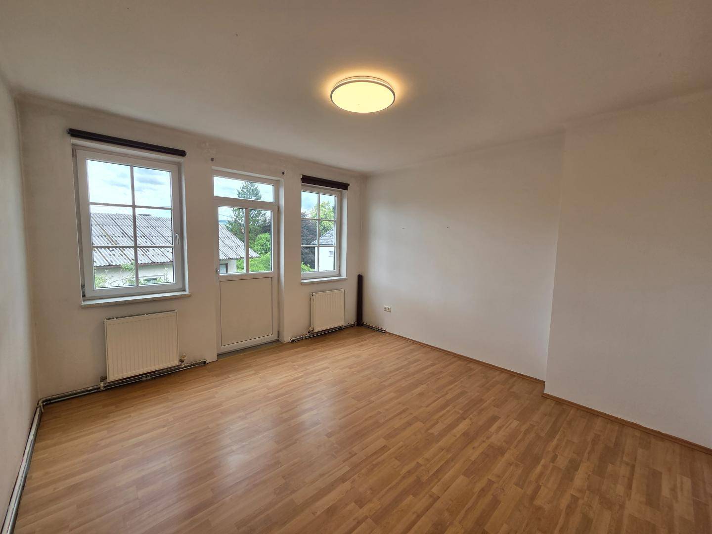 Leeres Zimmer mit Holzboden, zwei Fenstern und einer Tür, die nach draußen führt.