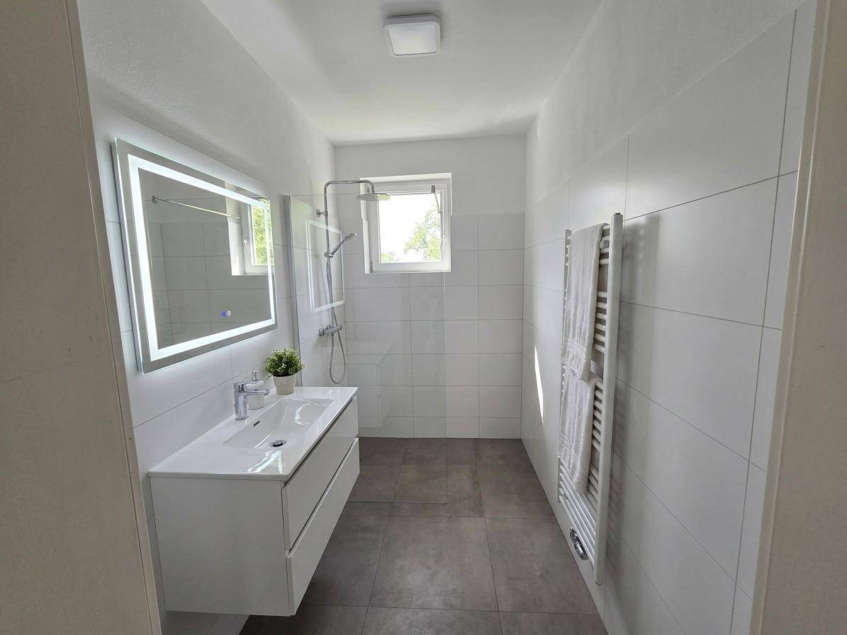 Modernes Badezimmer mit Dusche, Waschtisch und beleuchtetem Spiegel, hell gefliest.