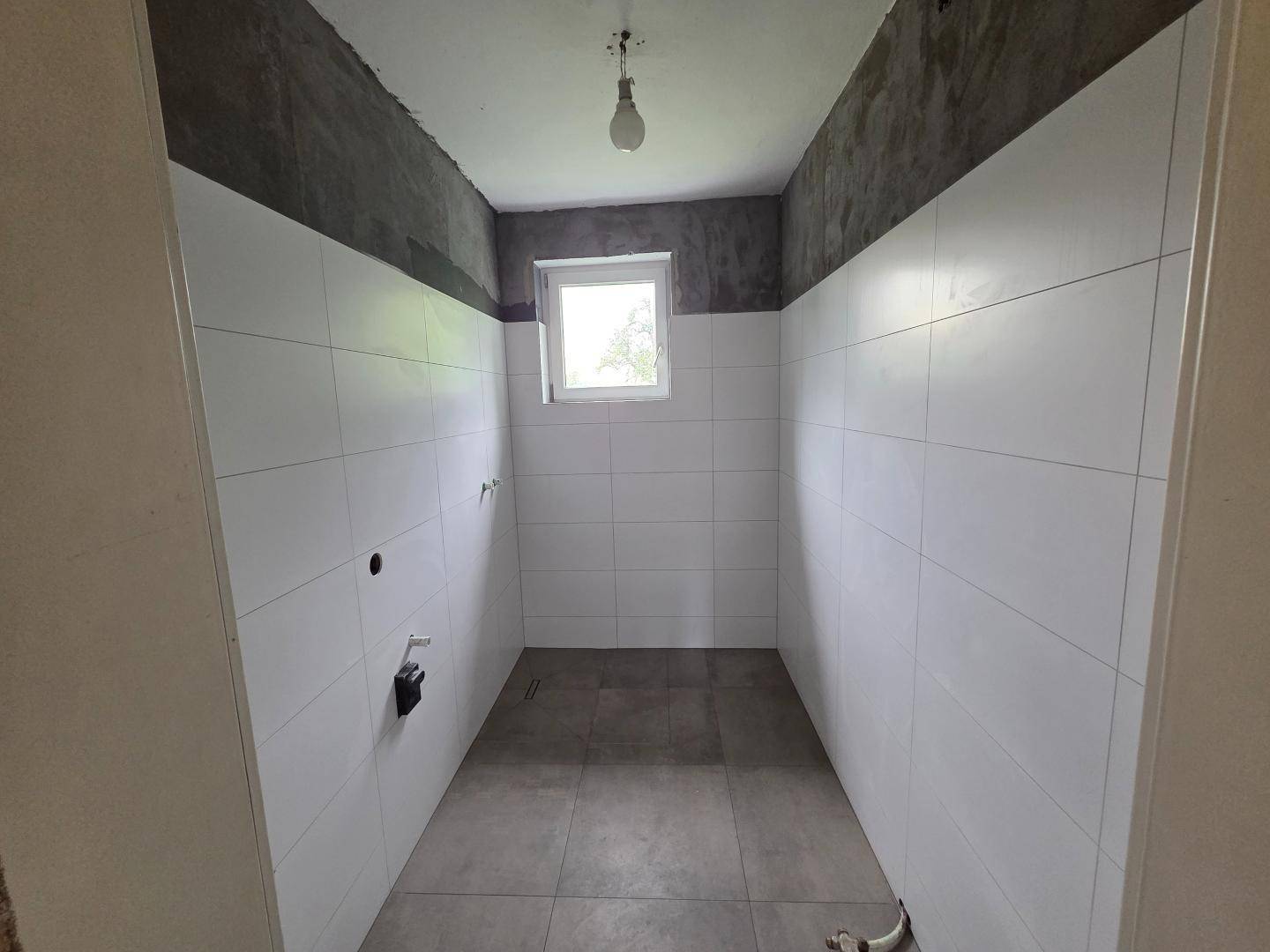 Badezimmer im Rohbau mit weißen Fliesen und Fenster, bereit für die Fertigstellung.