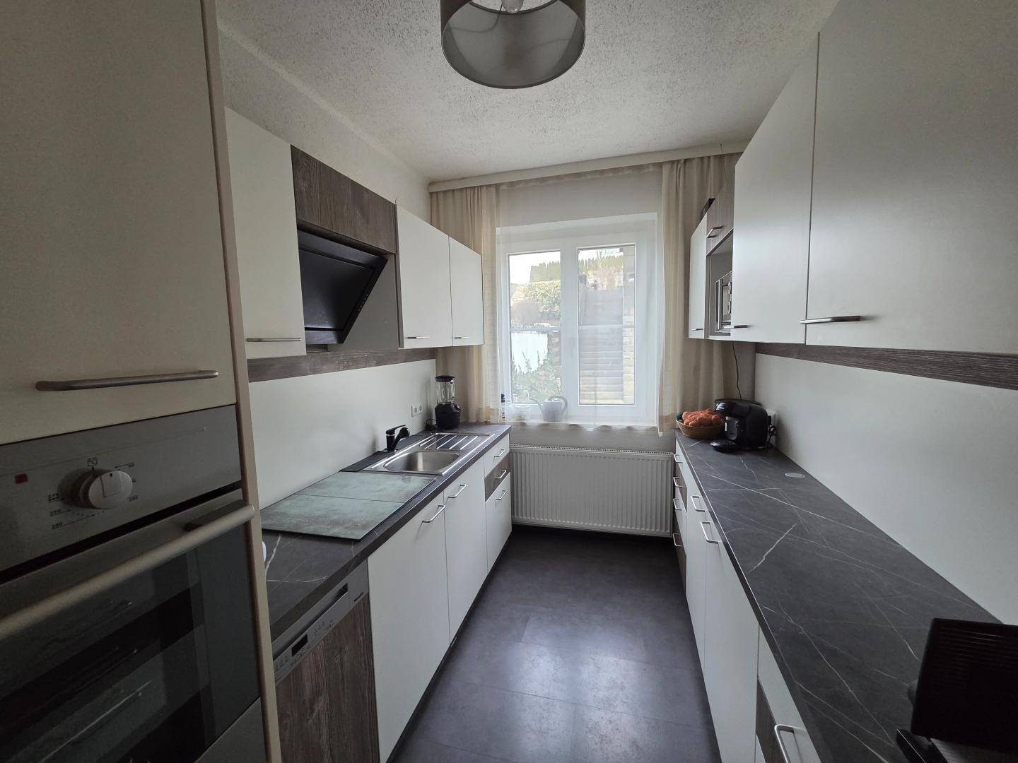 Moderne Einbauküche mit weißen Schränken, dunkler Arbeitsplatte und Fensterblick.