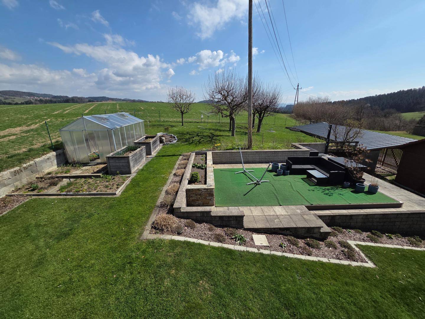 Großer Gartenbereich mit Gewächshaus, Hochbeeten und einer gepflasterten Sitzgelegenheit mit Weitblick.