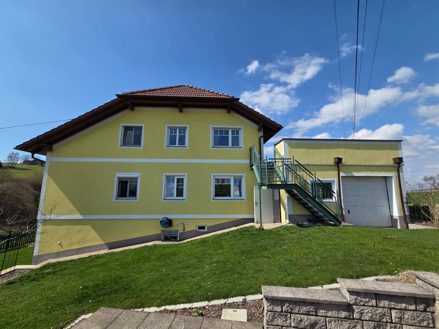 Rückansicht des gelben Hauses mit grünem Rasen und einer Außentreppe unter blauem Himmel.