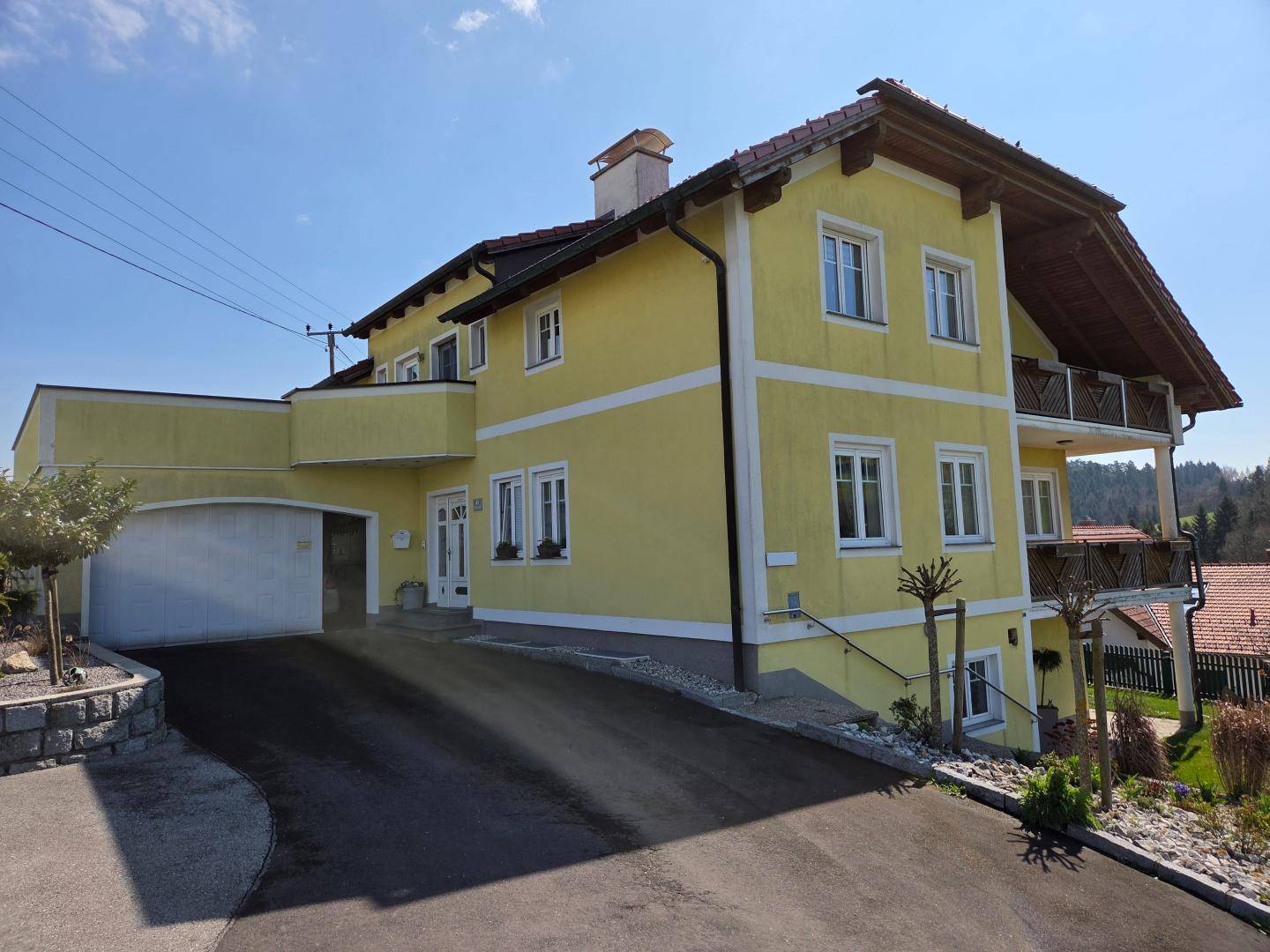 Seitenansicht des gelben Hauses mit Garage, Auffahrt und Balkonen in ländlicher Umgebung.