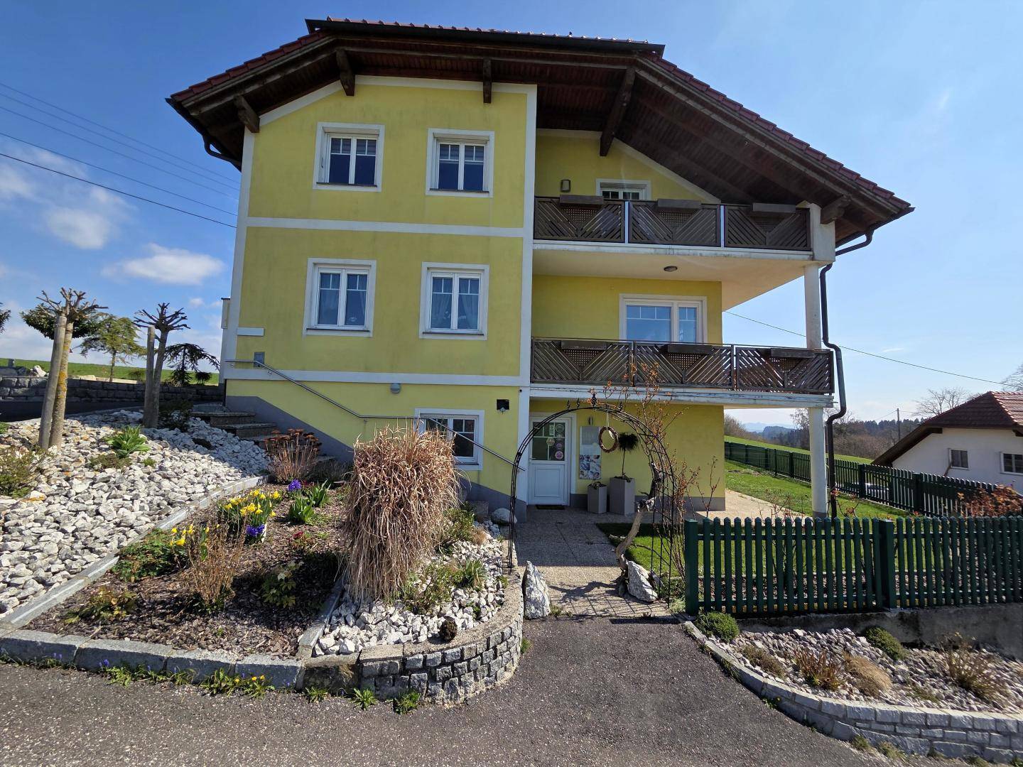 Gelbes Wohnhaus mit mehreren Balkonen und einem gepflegten Vorgarten unter blauem Himmel.