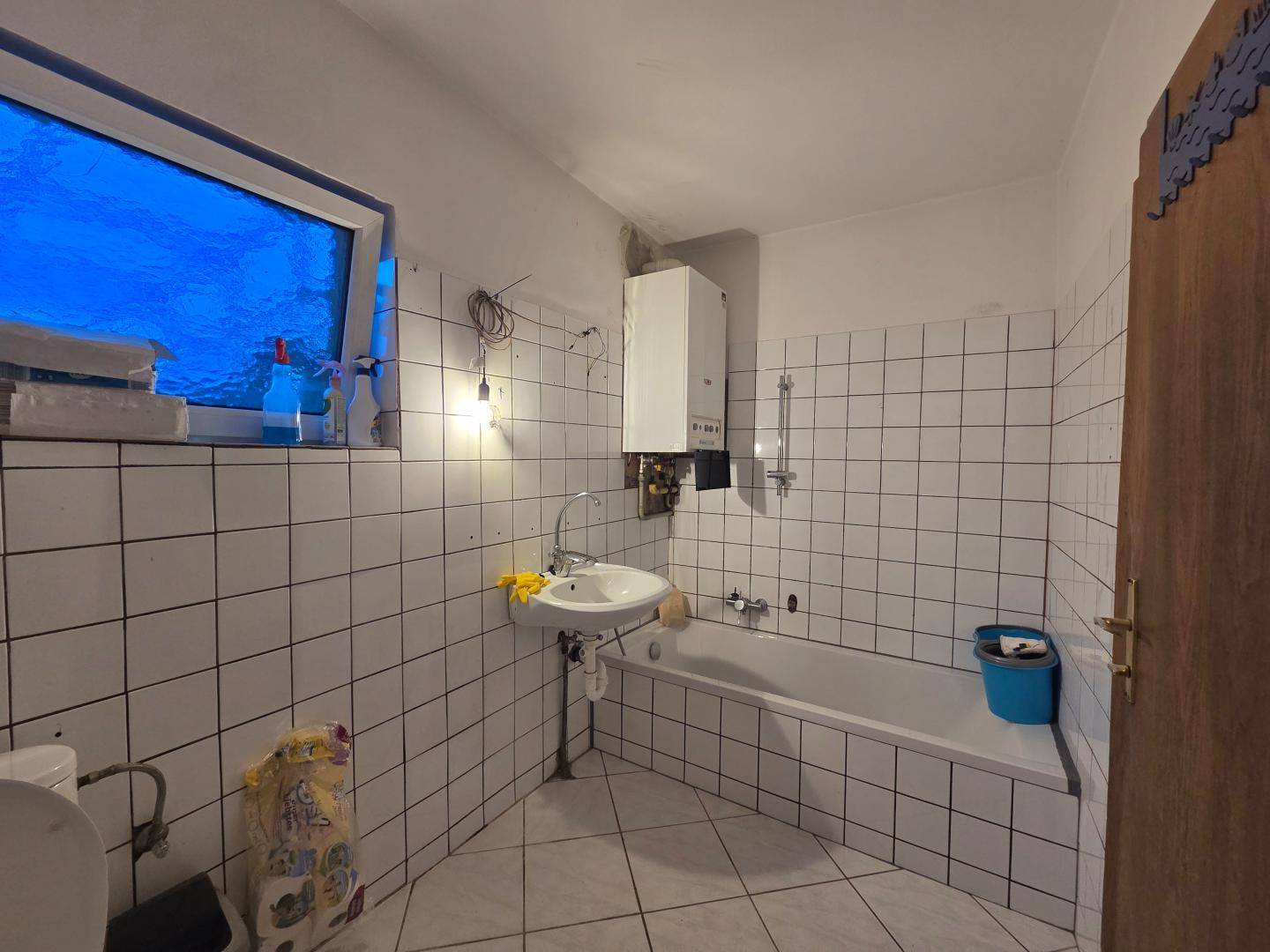 Älteres Badezimmer mit weißen Fliesen, Badewanne und einem Wandboiler, der renovierungsbedürftig ist.
