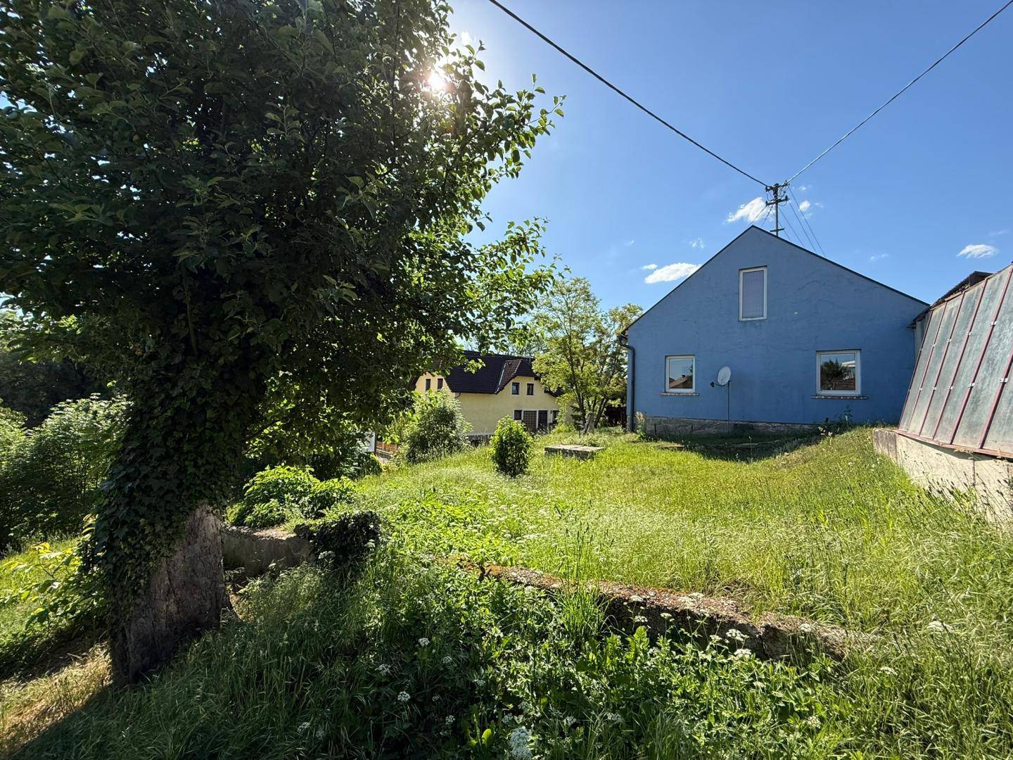 Blaues Haus mit angrenzendem Gewächshaus und weitem Blick ins Grüne an einem sonnigen Tag.