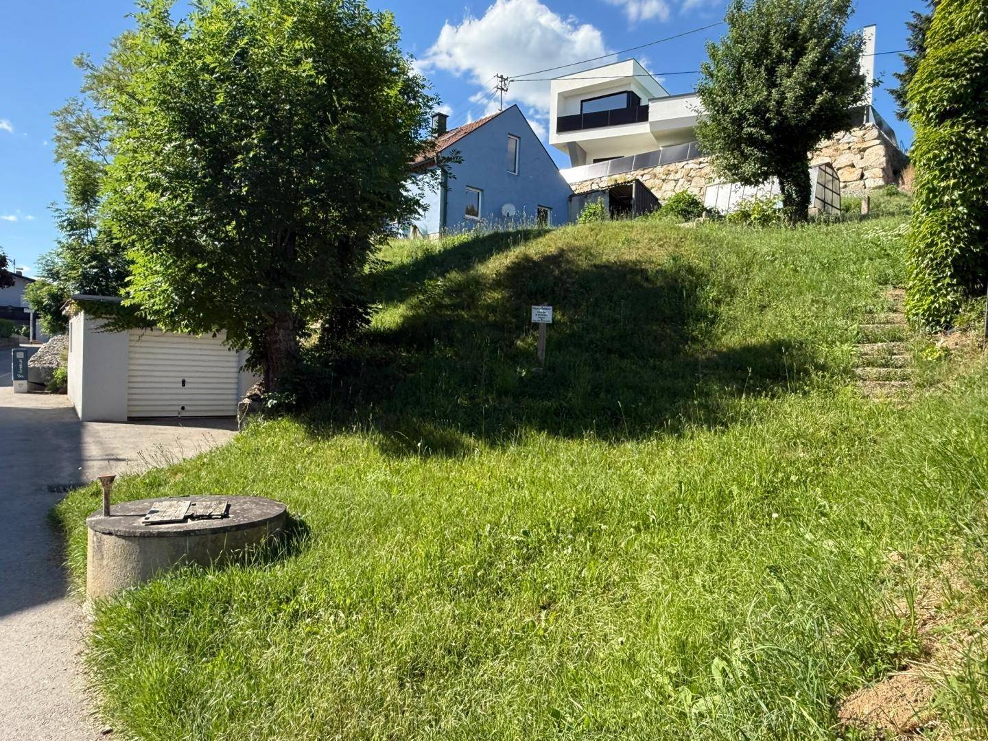 Grünes Grundstück mit einem blauen Haus und einem modernen Gebäude im Hintergrund unter blauem Himmel.
