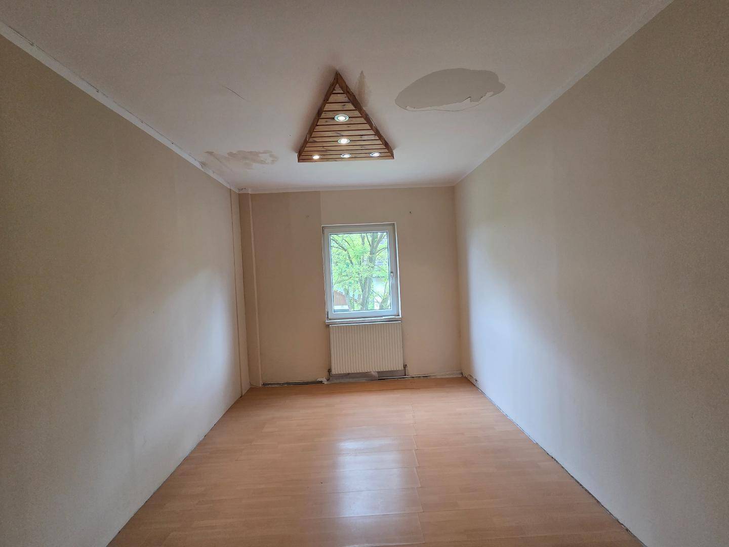 Helles Zimmer mit Laminatboden, Fenster und einer dreieckigen Holzdekorleuchte an der Decke.