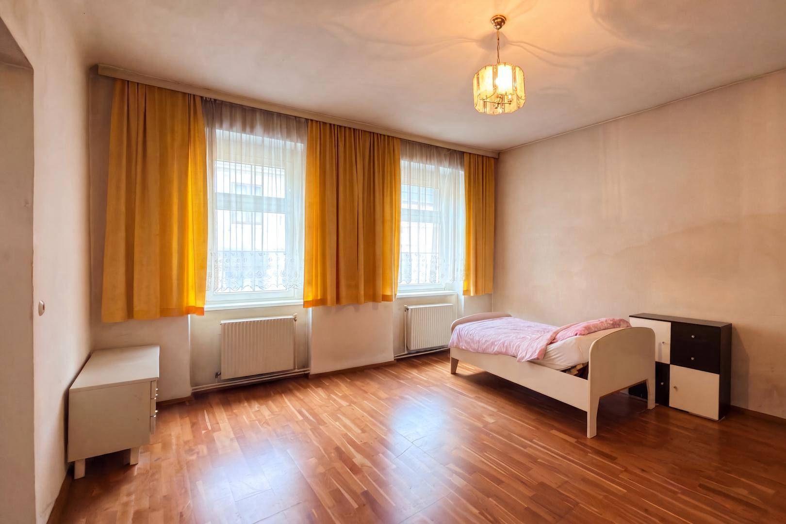 Schlafzimmer in ursprünglichem Zustand mit Holzboden, gelben Vorhängen und einfacher Einrichtung.