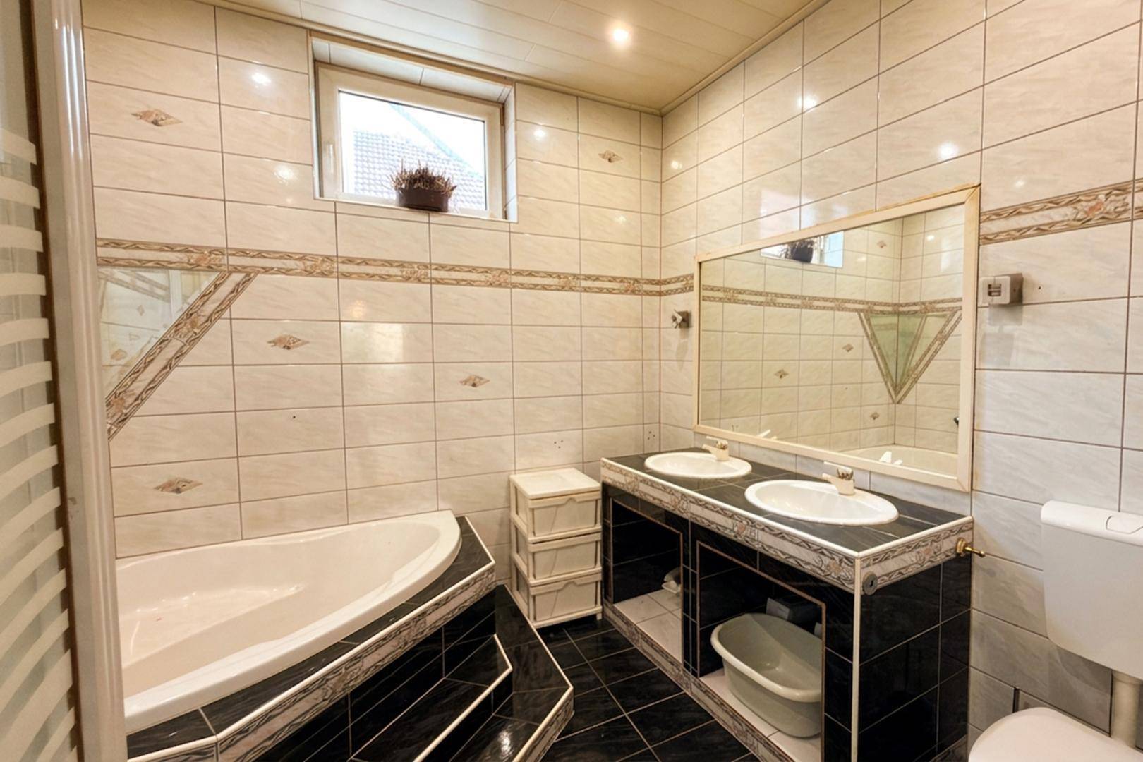 Badezimmer mit Fliesen im 80er-Jahre-Stil, Eckbadewanne, Doppelwaschbecken und einem Fenster.