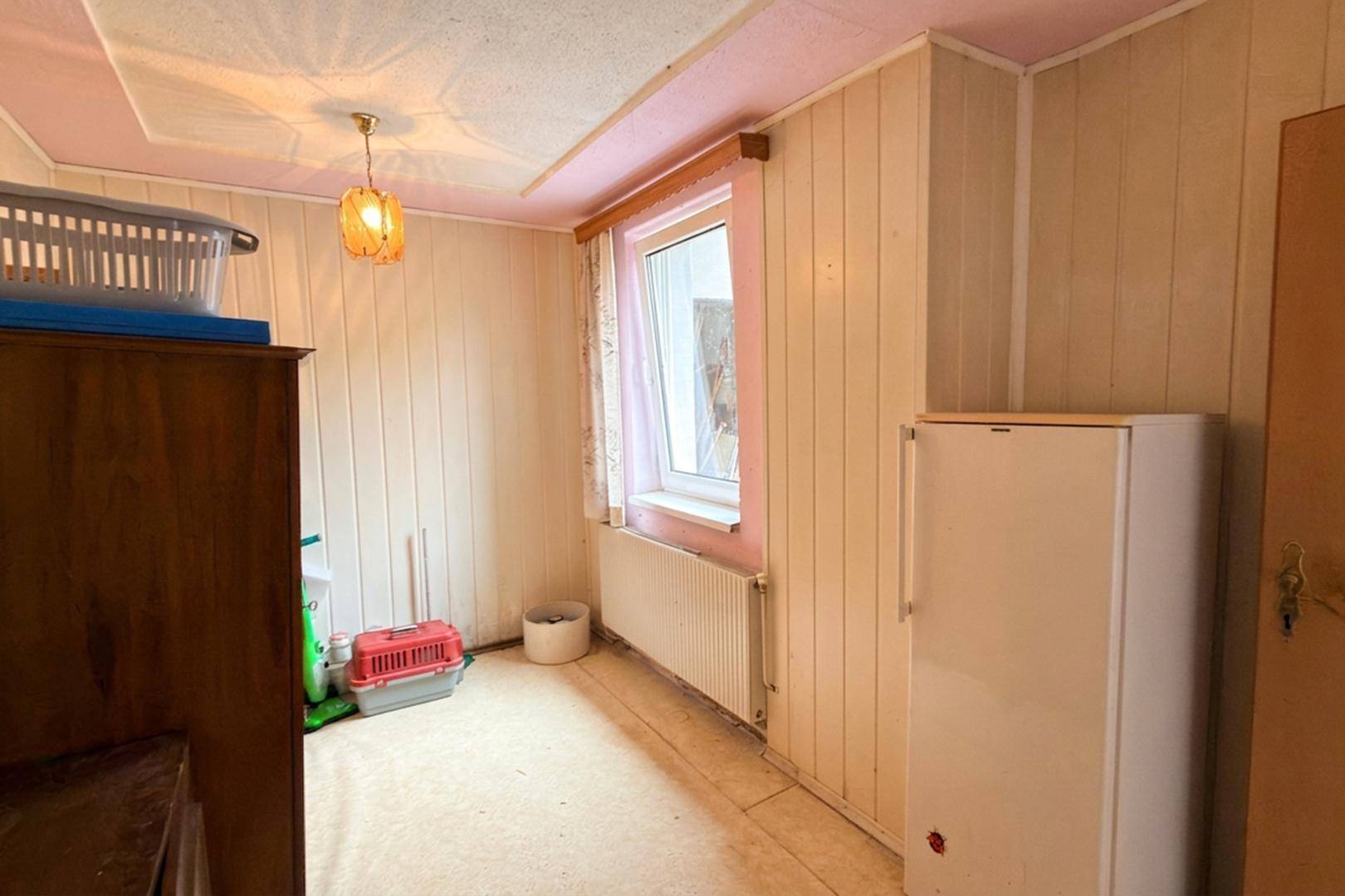 Kleiner Raum mit Holzpaneelen an den Wänden, rosa Decke und einem Fenster, das Tageslicht hereinlässt.