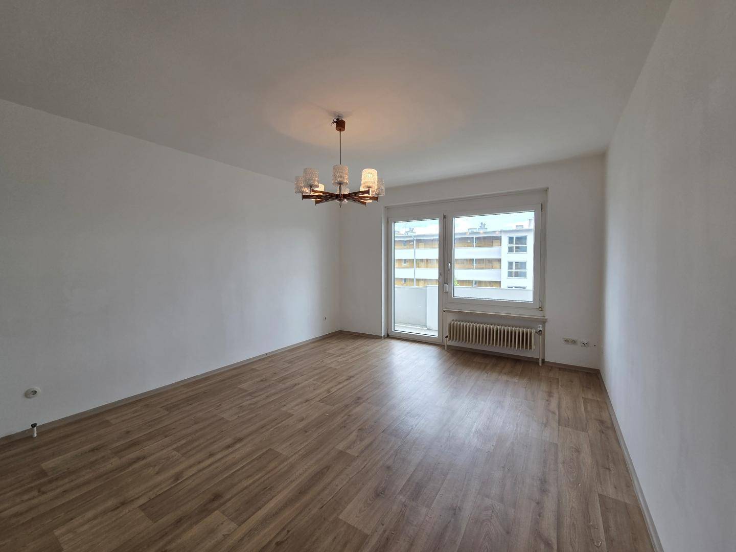 Helles, leeres Wohnzimmer mit Laminatboden, großem Fenster und Zugang zu einem Balkon.