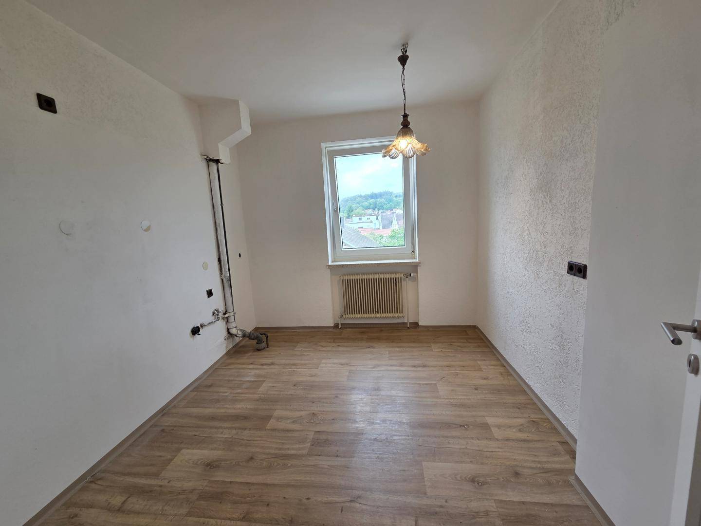Leerer Raum, vorbereitet für eine Küche, mit Fenster, Heizkörper und sichtbaren Anschlüssen.