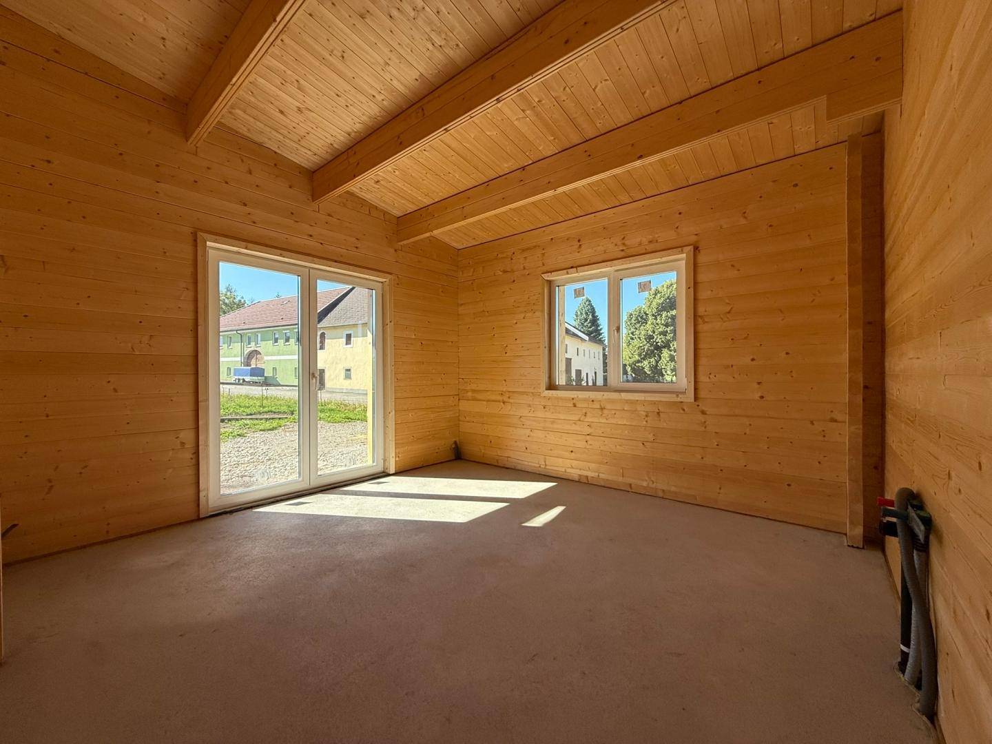 Unfertiger Raum mit Holzwänden, sichtbaren Deckenbalken und einem großen Fenster sowie einem kleineren Fenster.