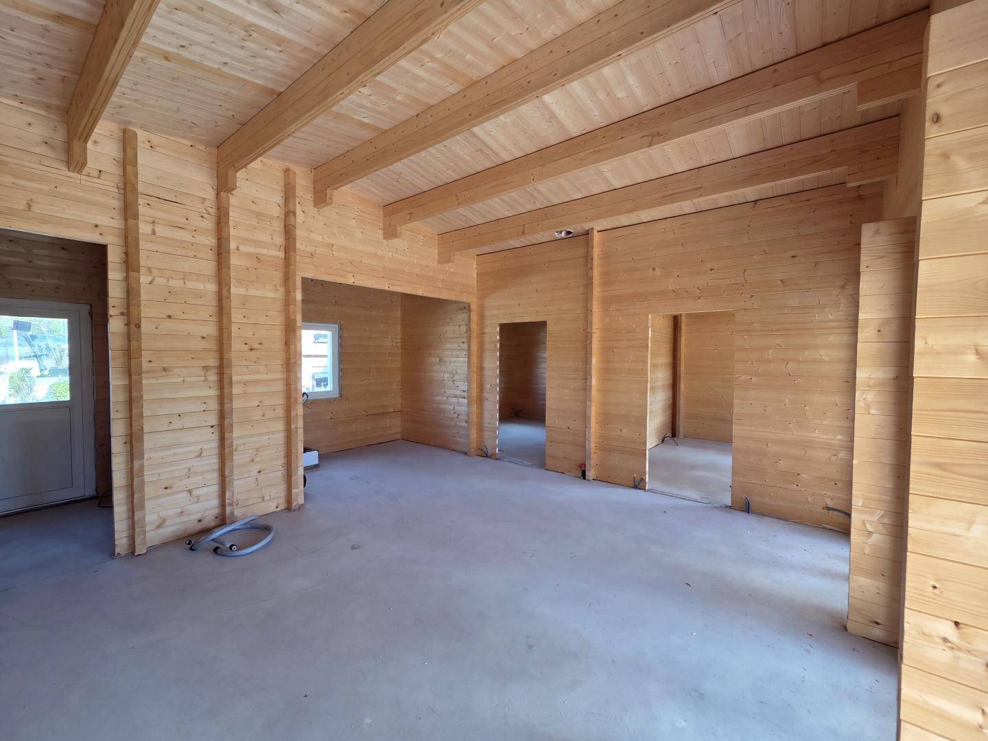 Unfertiger Innenraum eines Holzhauses mit sichtbaren Holzbalken und unbehandeltem Betonboden.