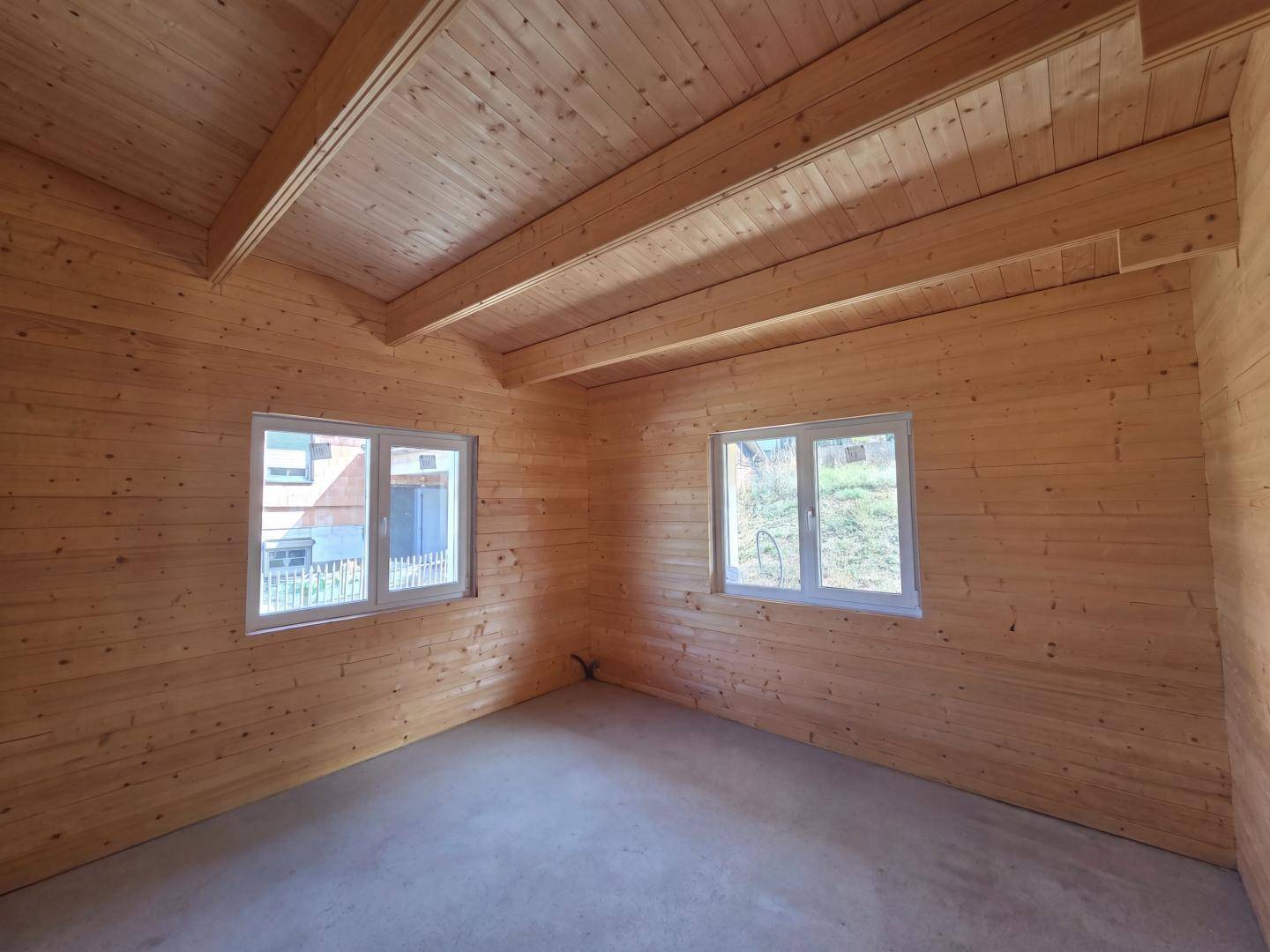 Unfertiger Raum mit Holzwänden, sichtbaren Deckenbalken und zwei Fenstern, die Tageslicht spenden.