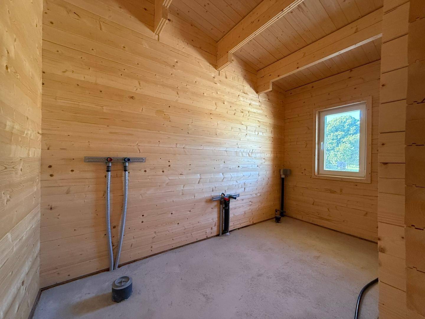 Unfertiger Raum mit Holzwänden, sichtbaren Deckenbalken und Anschlüssen für Sanitärinstallationen.