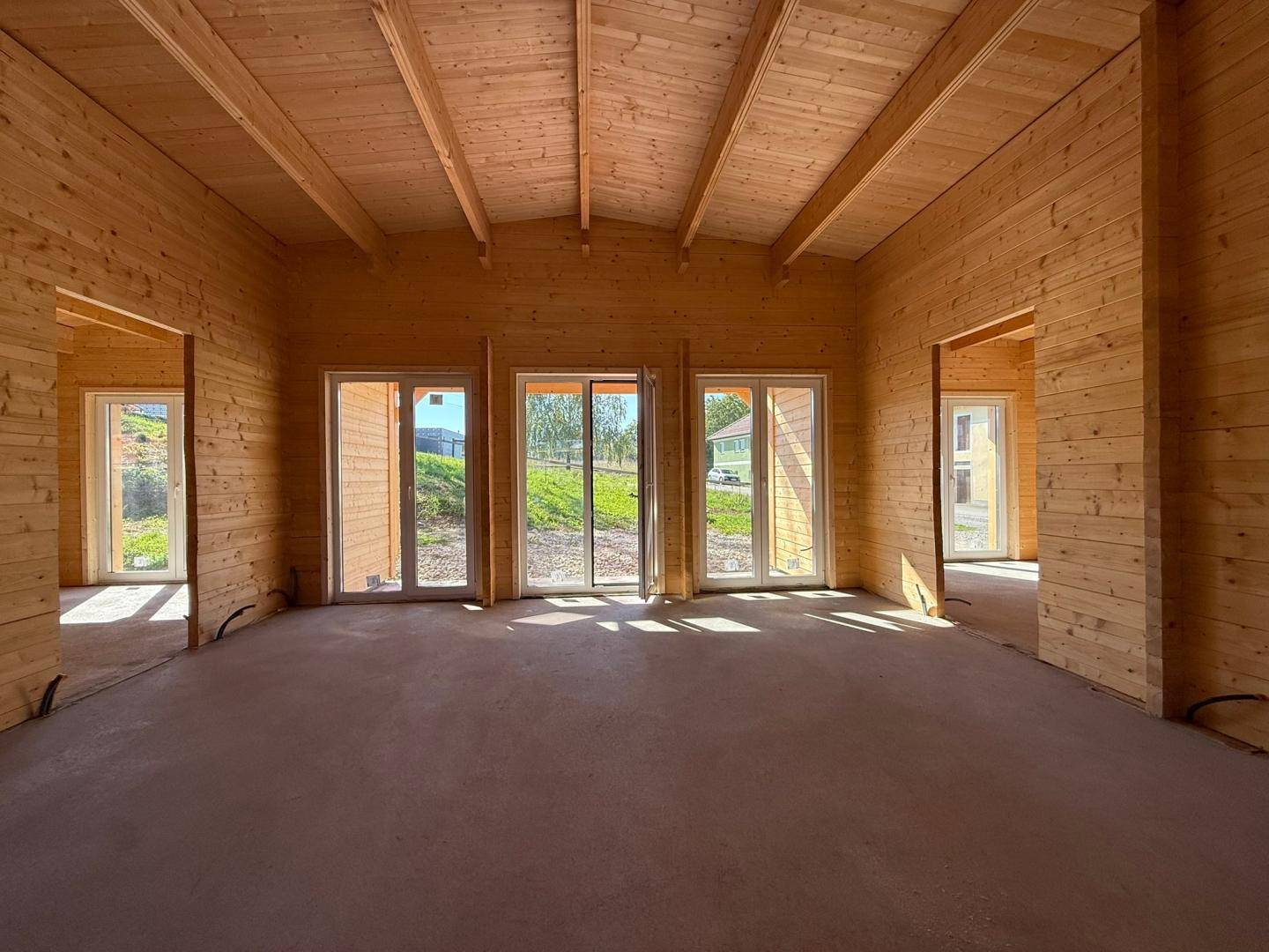 Unfertiger Innenraum mit Holzwänden, sichtbaren Deckenbalken und großen Fenstern mit Blick ins Grüne.