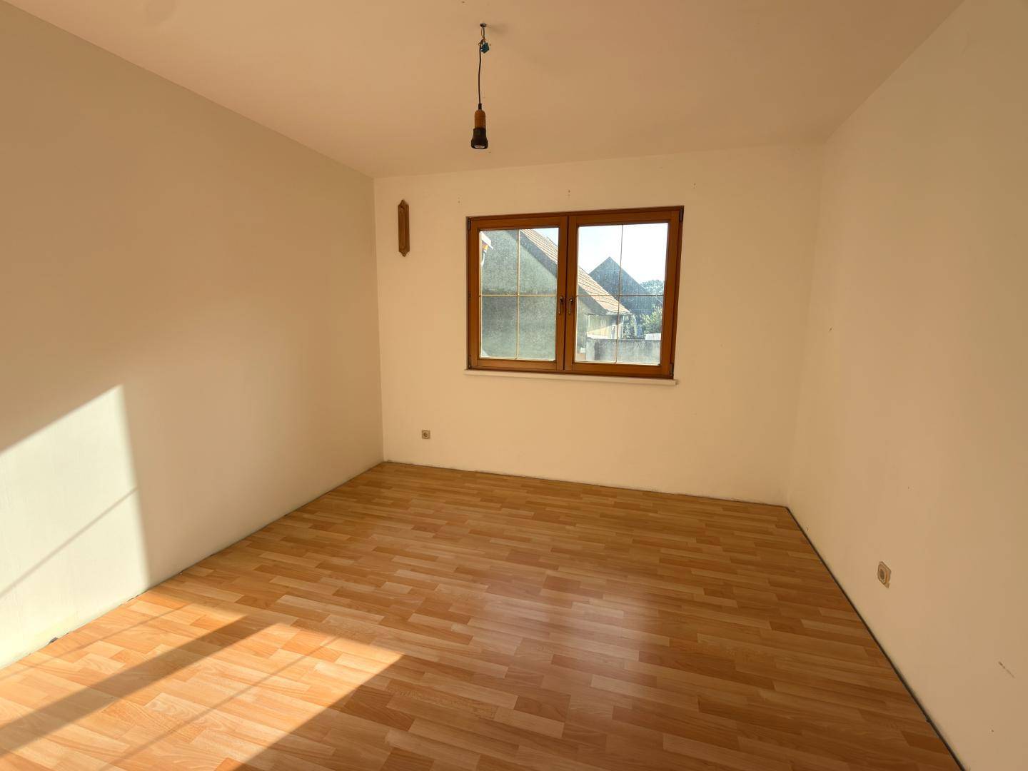 Leeres Schlafzimmer mit hellem Laminatboden und Fenster, das viel Licht hereinlässt.