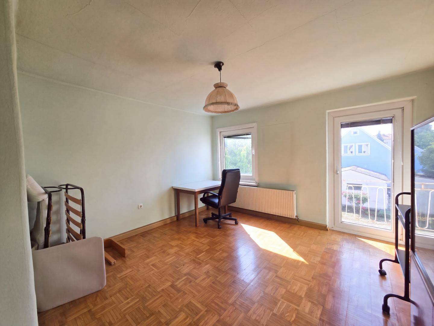 Schlafzimmer mit Parkettboden, Schreibtisch und Zugang zu einem kleinen Balkon.