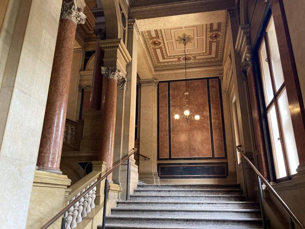 Elegantes Treppenhaus mit Marmorstufen, roten Marmorsäulen und einer verzierten Decke mit Kronleuchter.