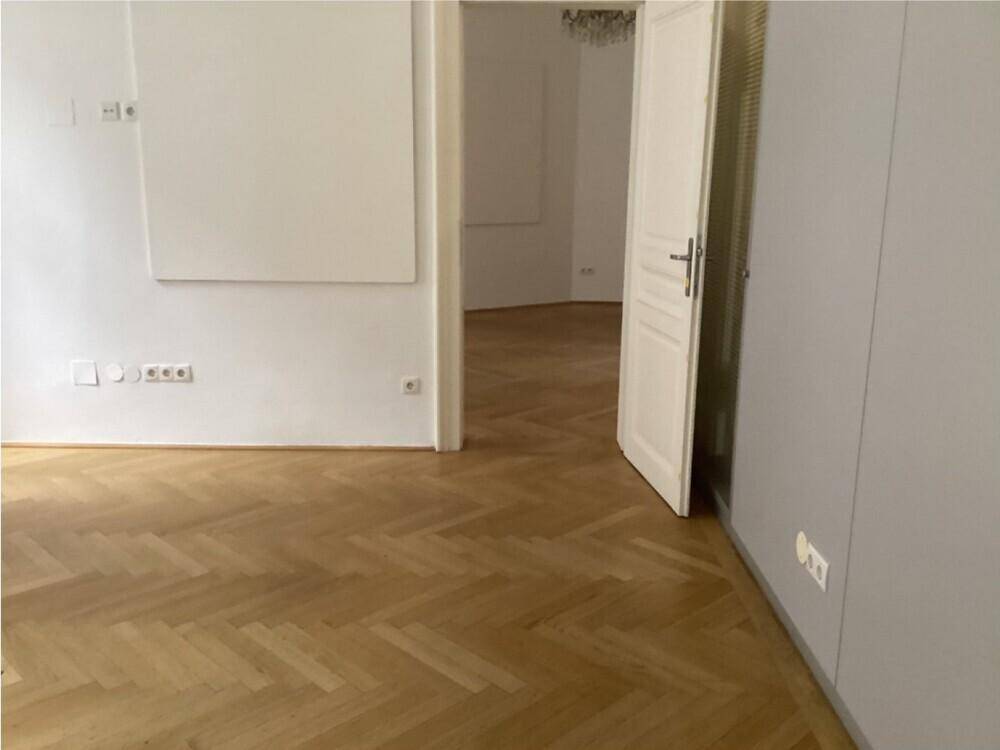 Helles Zimmer mit Fischgrätparkett, weißen Wänden und einer Tür zu einem weiteren Raum.