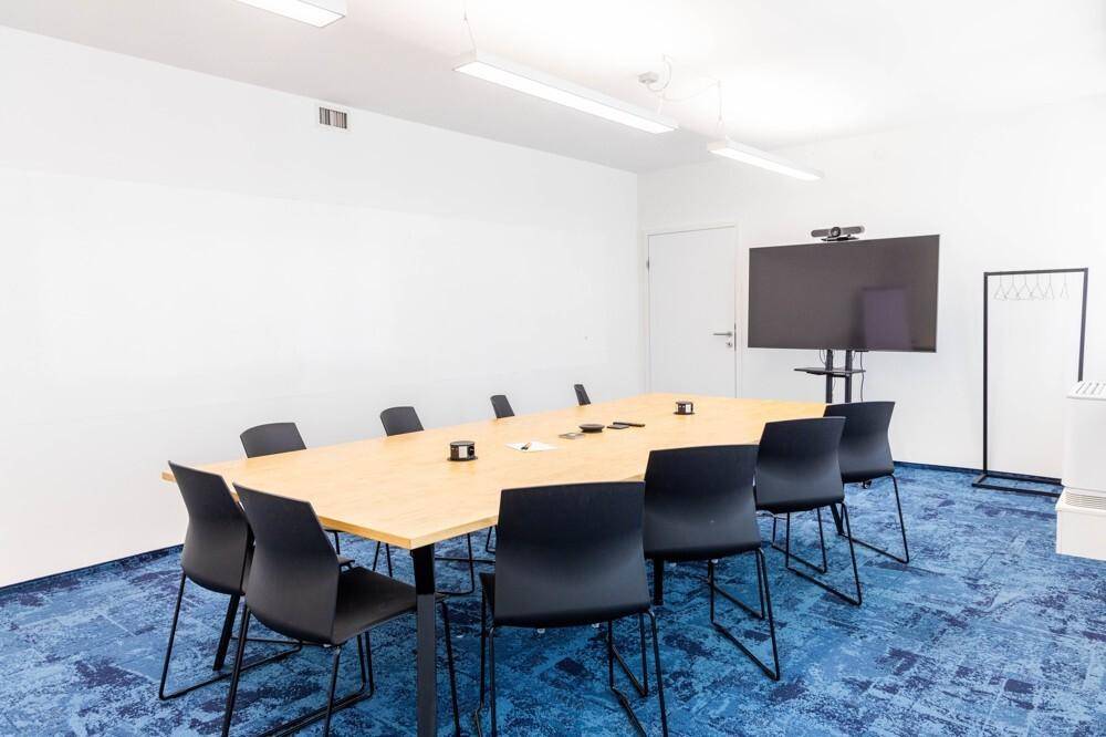 Heller Konferenzraum mit großem Holztisch, blauer Teppichboden und einem großen Bildschirm für Präsentationen.