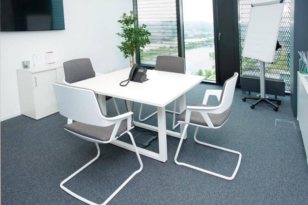 Kleiner Besprechungsraum mit quadratischem Tisch, modernen Stühlen und einem Flipchart für Präsentationen.
