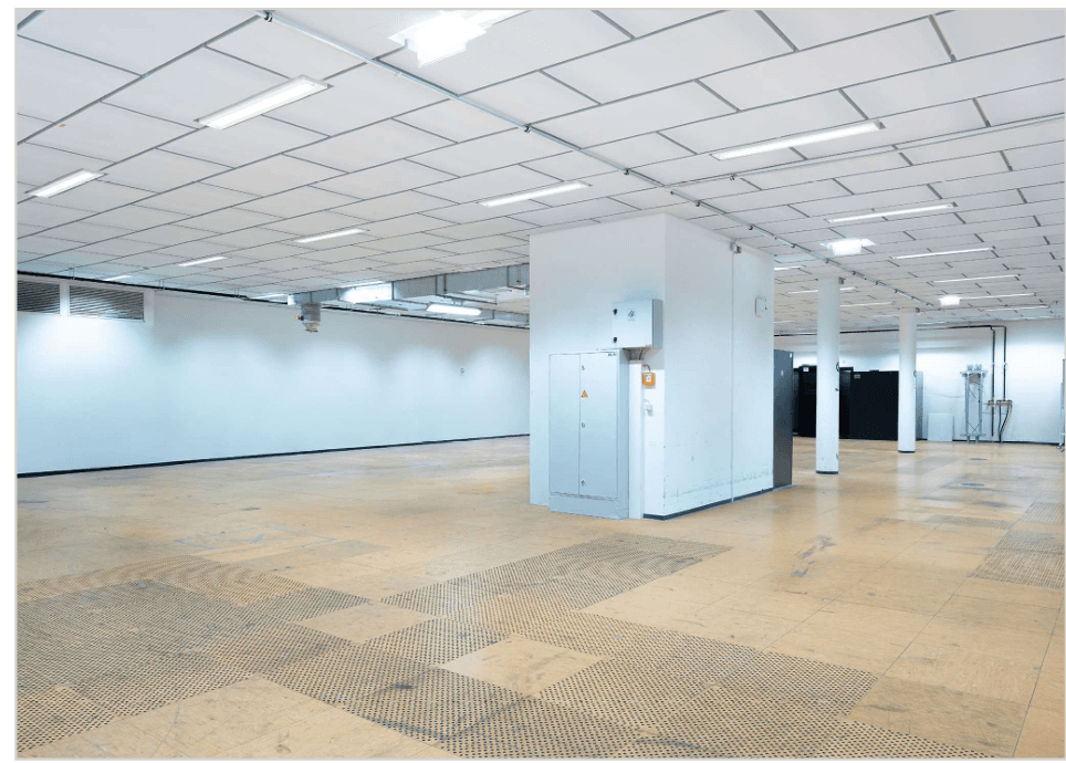 Geräumiger, leerer Gewerberaum mit erhöhtem Boden, sichtbaren Deckeninstallationen und einem zentralen Technikbereich.