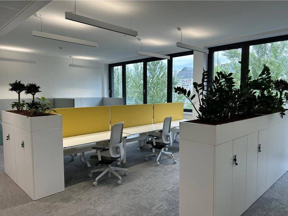 Heller Großraumbüro-Arbeitsplatz mit modernen Schreibtischen, ergonomischen Stühlen und Pflanzen als Raumteiler.