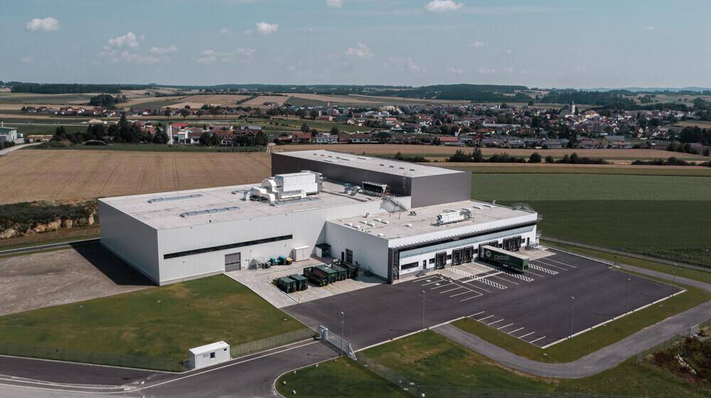 Weitläufige Luftaufnahme eines modernen Industriegebäudes mit angrenzendem Dorf und Feldern.